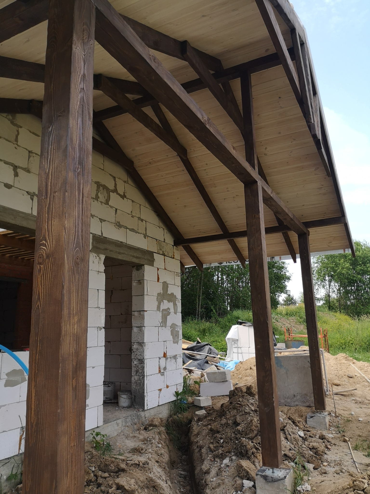 Surowy stan budowy zadaszenia tarasu: drewniane słupy z belek, białe bloczki ścian, widoczny wykop w ziemi i materiały budowlane w tle.