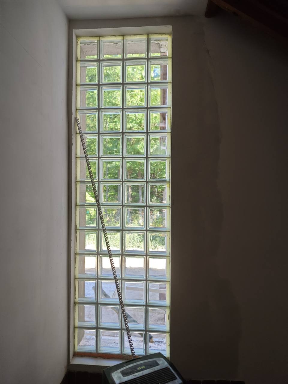 Wysoki, wąski świetlik z luksferów w surowym wnętrzu, widoczna drabina oparta o szybę i stary grzejnik elektryczny u dołu