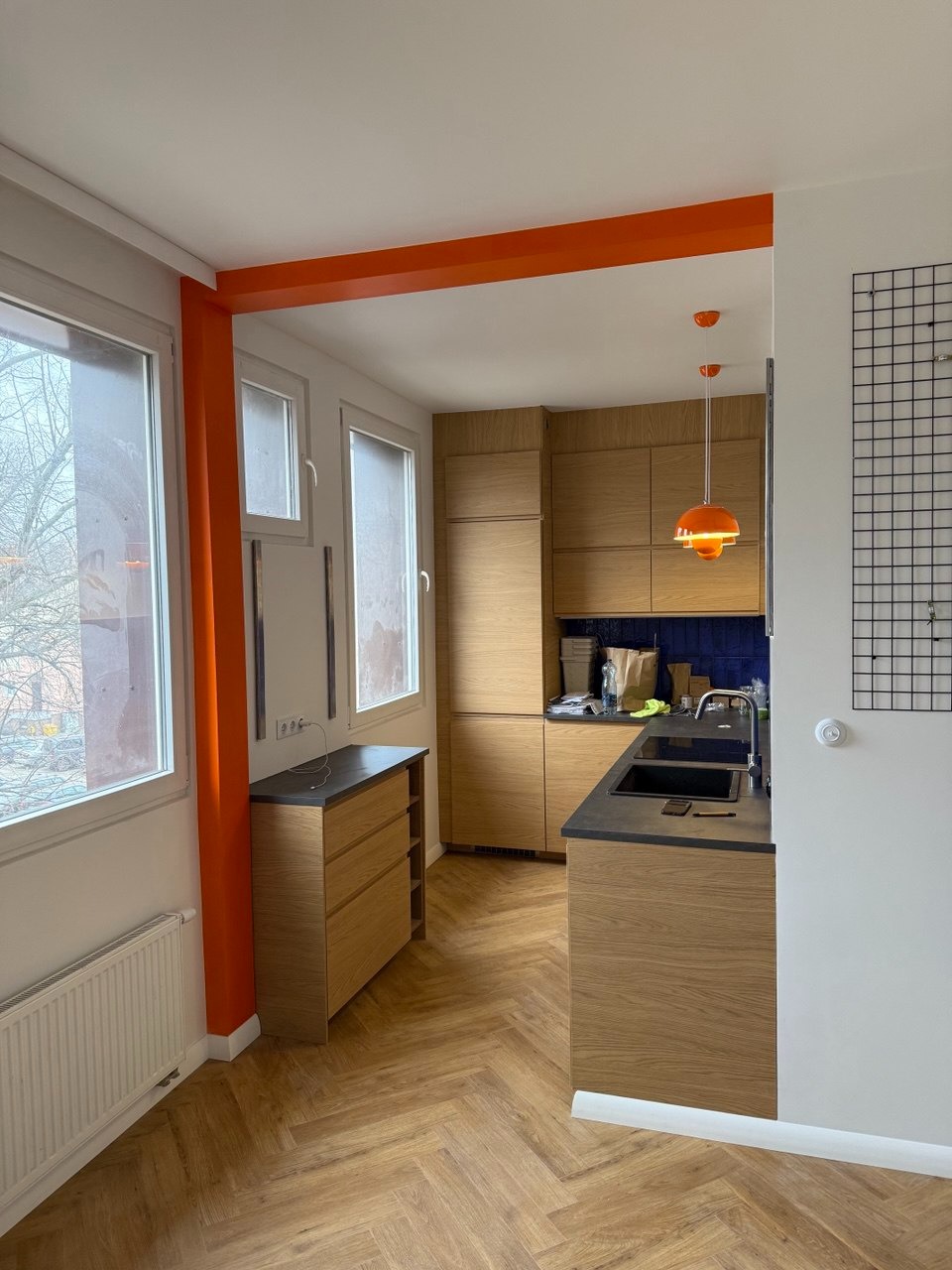 Nowoczesna kuchnia z drewnianymi szafkami i blatem w kolorze grafitu, parkiet ułożony w jodełkę, pomarańczowe akcenty na ścianach i lampach, widok z perspektywy wejścia.