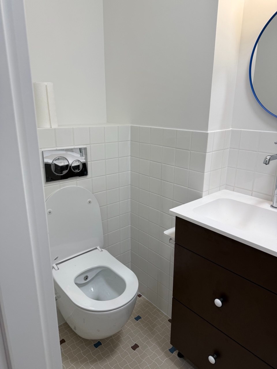 Nowoczesna toaleta z deską myjącą i podwieszaną spłuczką, białe płytki na ścianach, ciemnobrązowa szafka pod umywalką z białym blatem, wzorzysta podłoga w łazience.