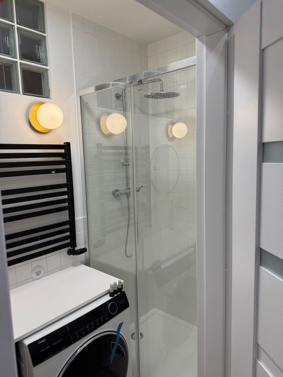 Biała łazienka z kabiną prysznicową, okrągłymi lampami ściennymi i pralką Haier z blatem.