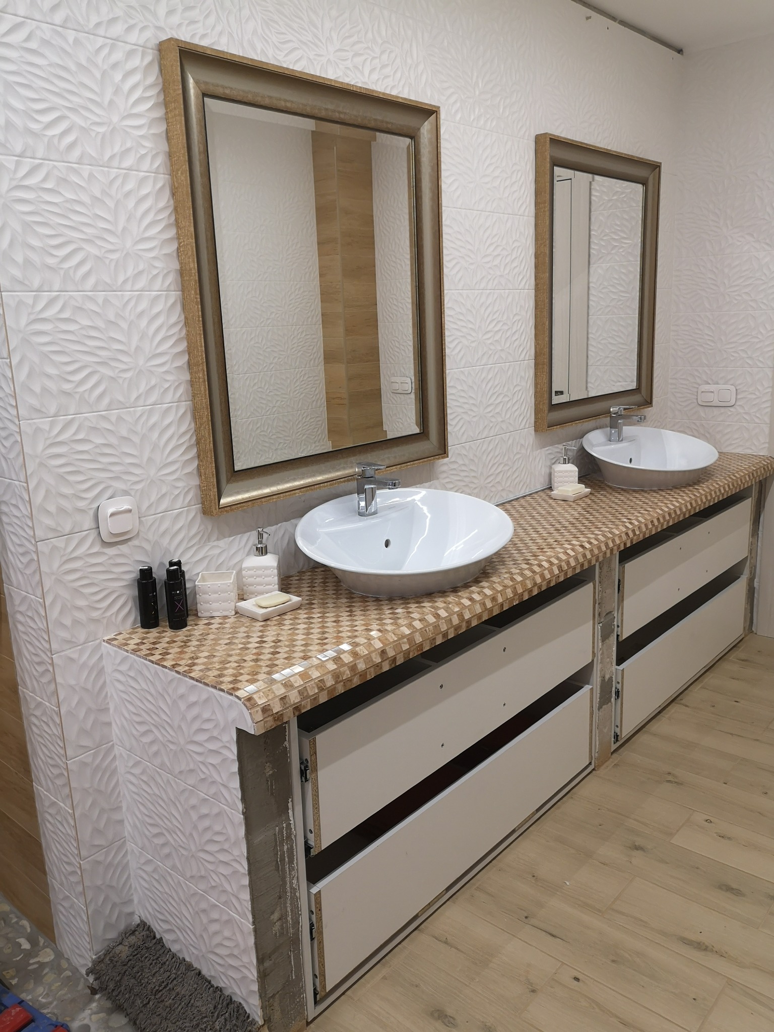 Dwie białe umywalki nablatowe z chromowanymi bateriami na drewnianym blacie z mozaiką, zawieszone nad szafkami z otwartymi szufladami, na tle ściany z białych płytek 3D i dwóch luster w złotej ramie.