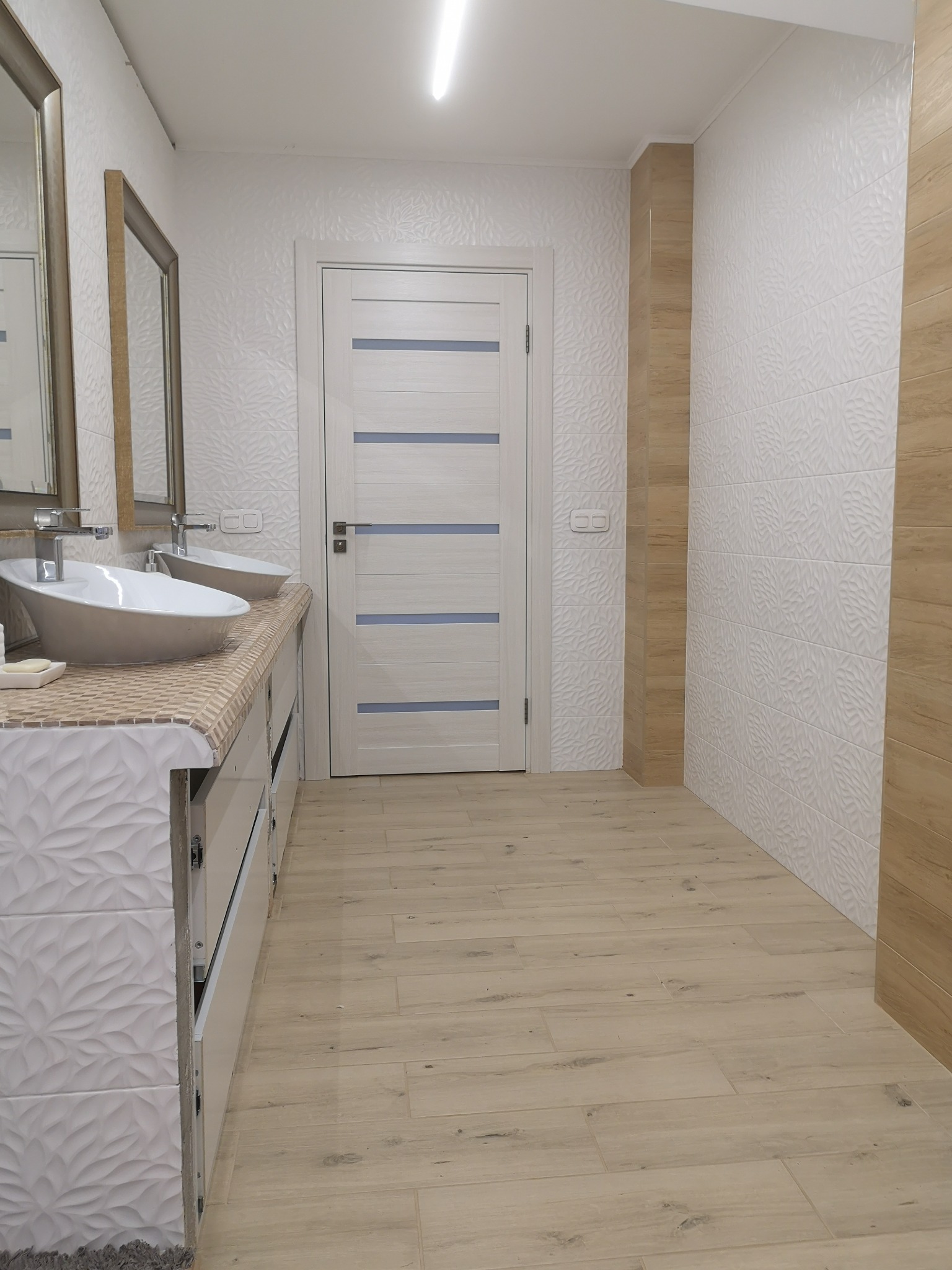 Jasna łazienka po remoncie z podwójną umywalką nablatową, mozaikowym blatem, białą teksturowaną ścianą, drewnopodobnymi płytkami podłogowymi i białą stolarką drzwiową z poziomymi paskami.