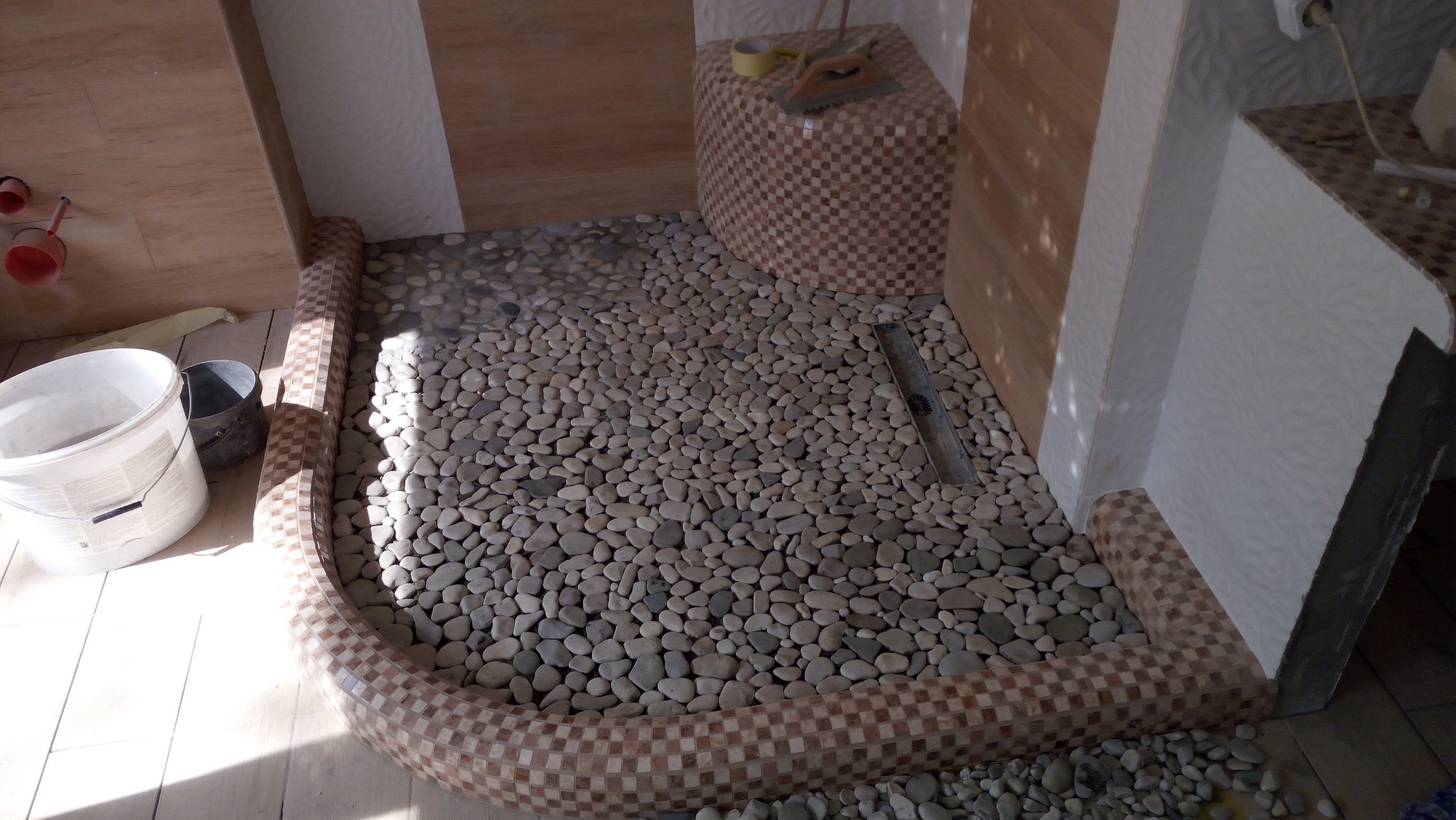 Prysznic w trakcie wykańczania z podłogą wyłożoną drobnymi kamieniami, obramowany mozaiką w brązowo-beżową szachownicę, widoczne białe i szare wiaderka, narzędzia na obudowie.