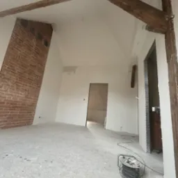 Pokój na poddaszu w trakcie remontu, ściany przygotowane pod malowanie, widoczne drewniane belki stropowe, fragment ściany z cegły, na podłodze rozłożona folia ochronna, zwinięty przedłużacz...