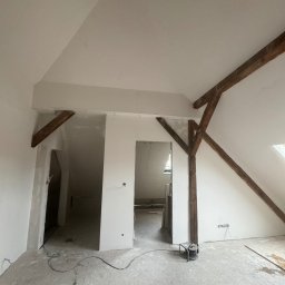 Jasiek Linkert - Poddasze po szpachlowaniu, widoczne drewniane elementy konstrukcyjne, ściany przygotowane do malowania, podłoga zabezpieczona folią. Widok z dołu, szeroki kąt.