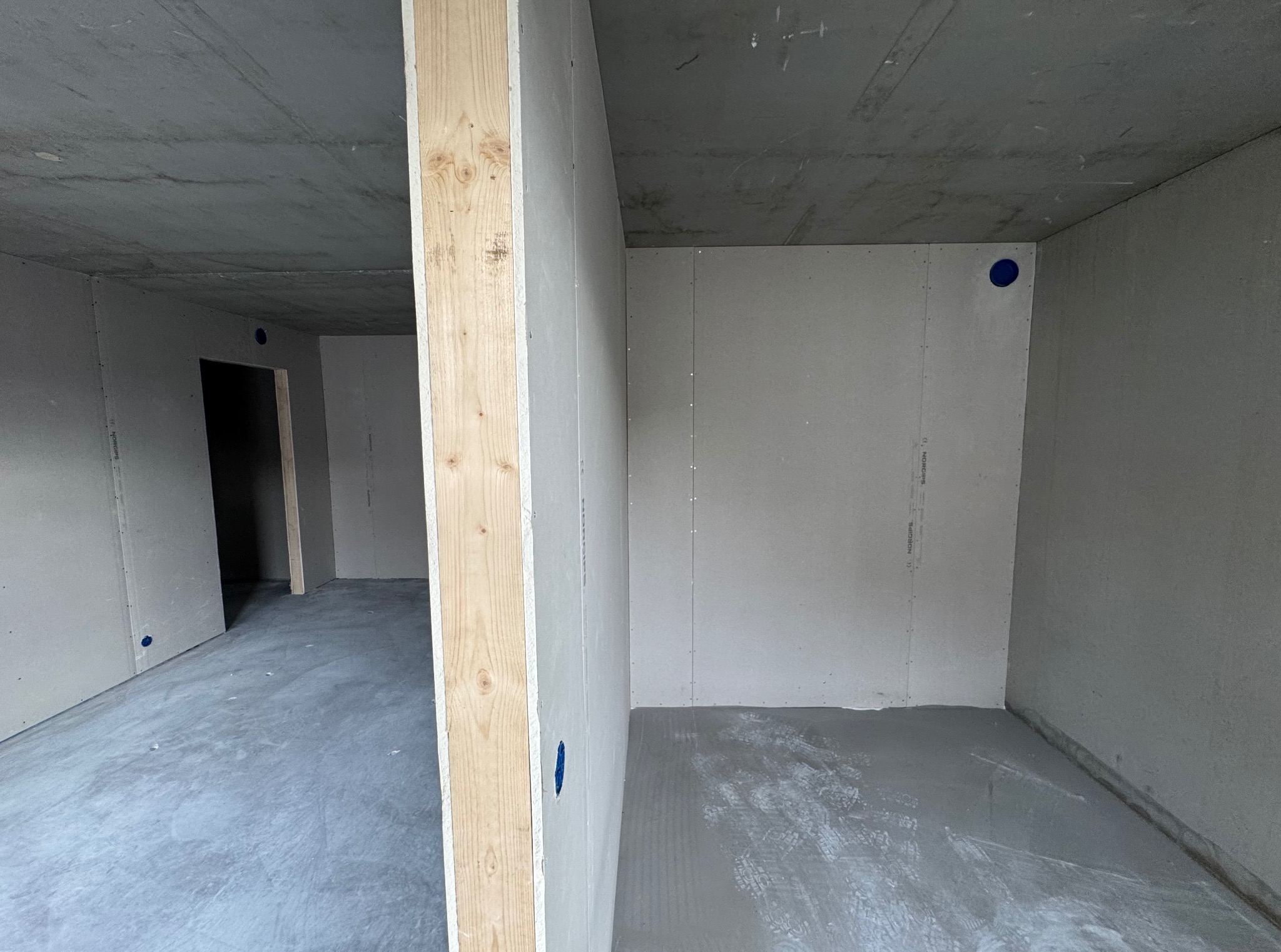 Surowe wnętrze w trakcie wykańczania: ściany z płyt, drewniane belki, szara podłoga. Widoczne niebieskie punkty instalacyjne. Minimalistyczny, industrialny klimat.