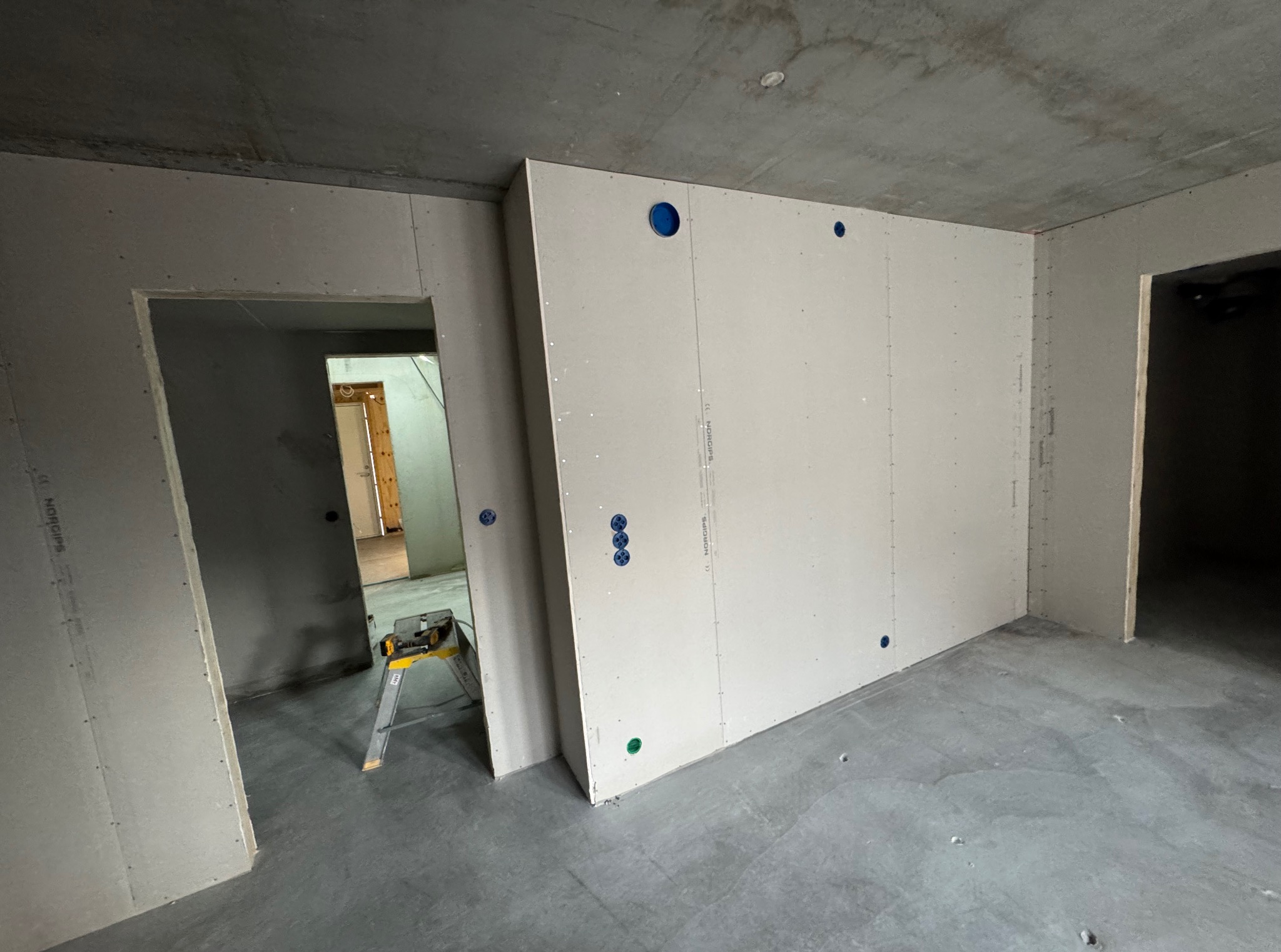 Wnętrze w trakcie prac wykończeniowych: ściany obłożone płytami gipsowo-kartonowymi z widocznymi otworami na instalacje elektryczne. W tle drabina malarska.