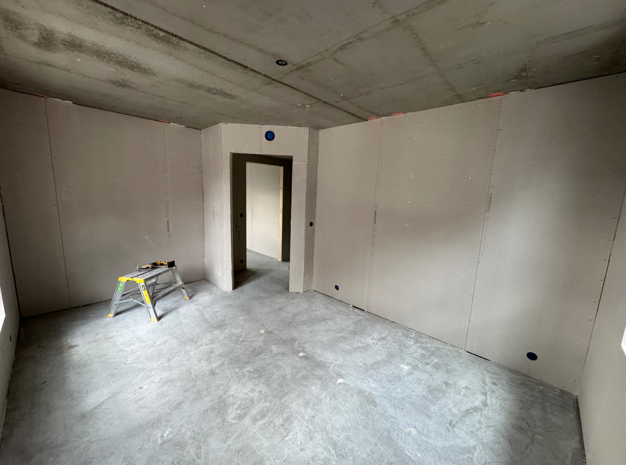 Pokój w trakcie remontu: ściany obłożone płytami, widoczna drabina malarska i surowy betonowy sufit z instalacją. Otwór drzwiowy w tle. Wnętrze przygotowane do dalszych prac wykończeniowych.