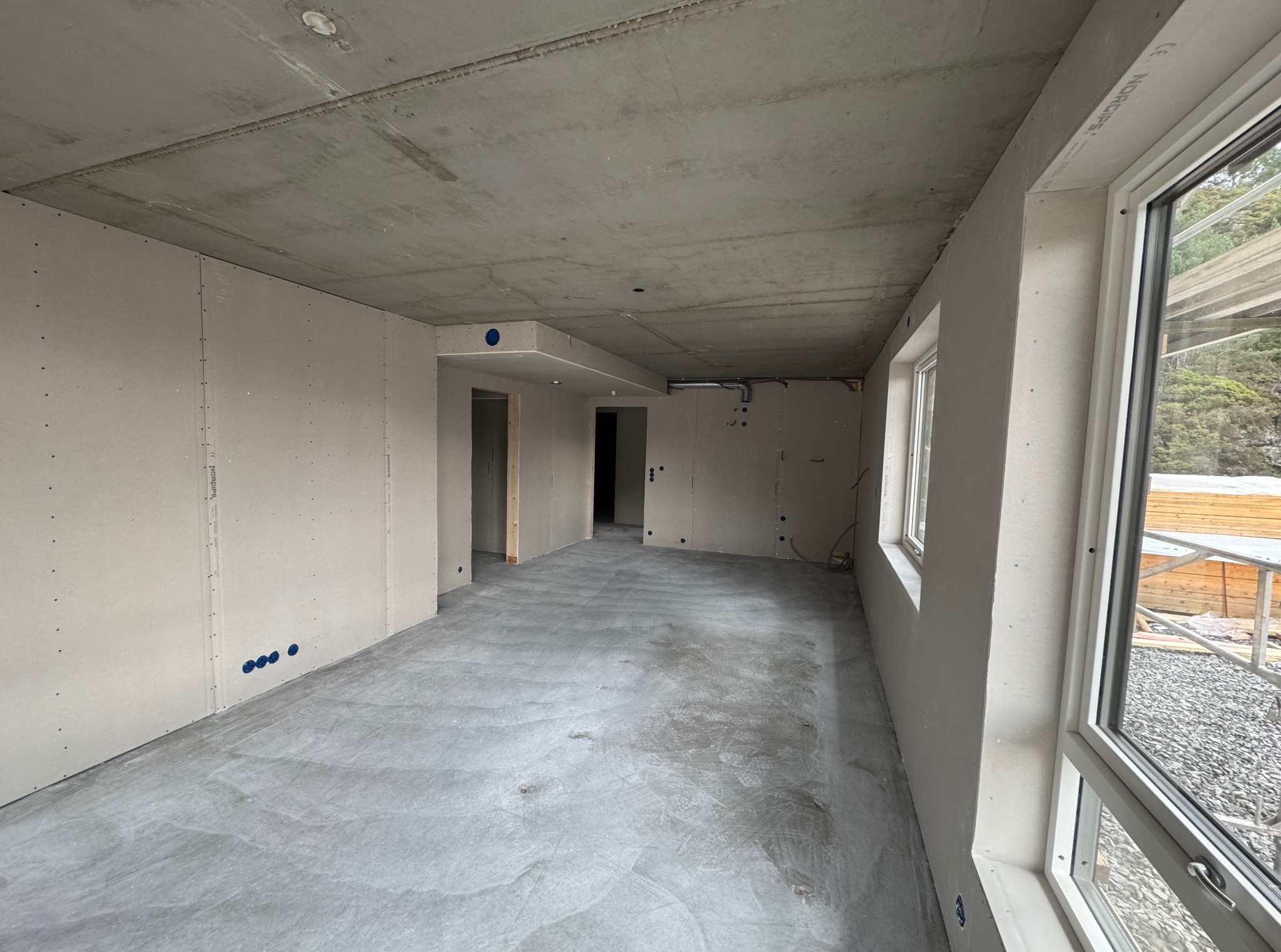 Wnętrze w trakcie remontu: surowy betonowy sufit, ściany wyłożone płytami, widoczne puszki elektryczne. Duże okno z widokiem na zewnątrz. Podłoga betonowa.