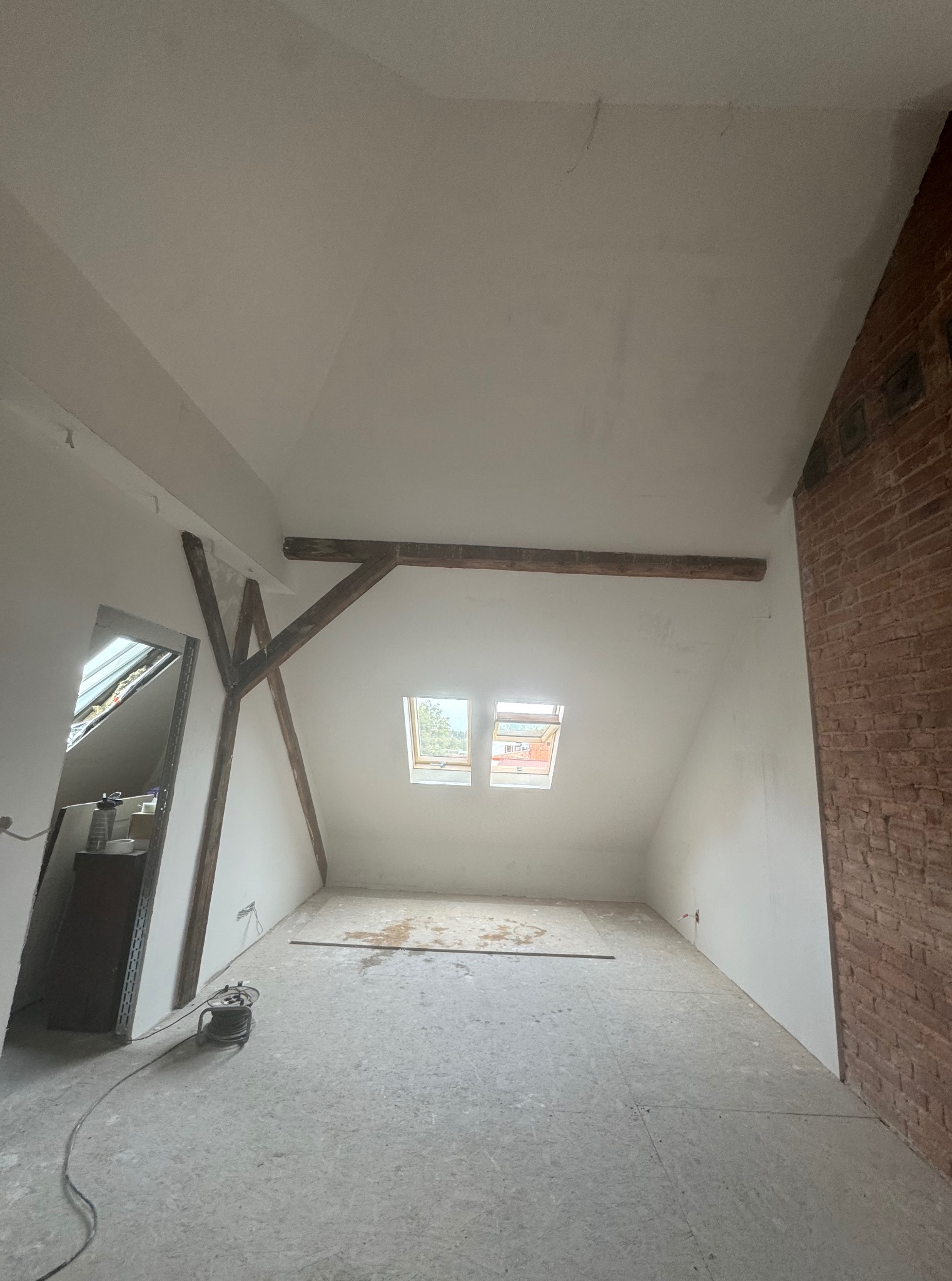 Poddasze po remoncie: białe ściany, drewniane belki, okno dachowe. Widoczna surowa podłoga i fragment ściany z cegły. Przy podłodze stoi zwinięty kabel.
