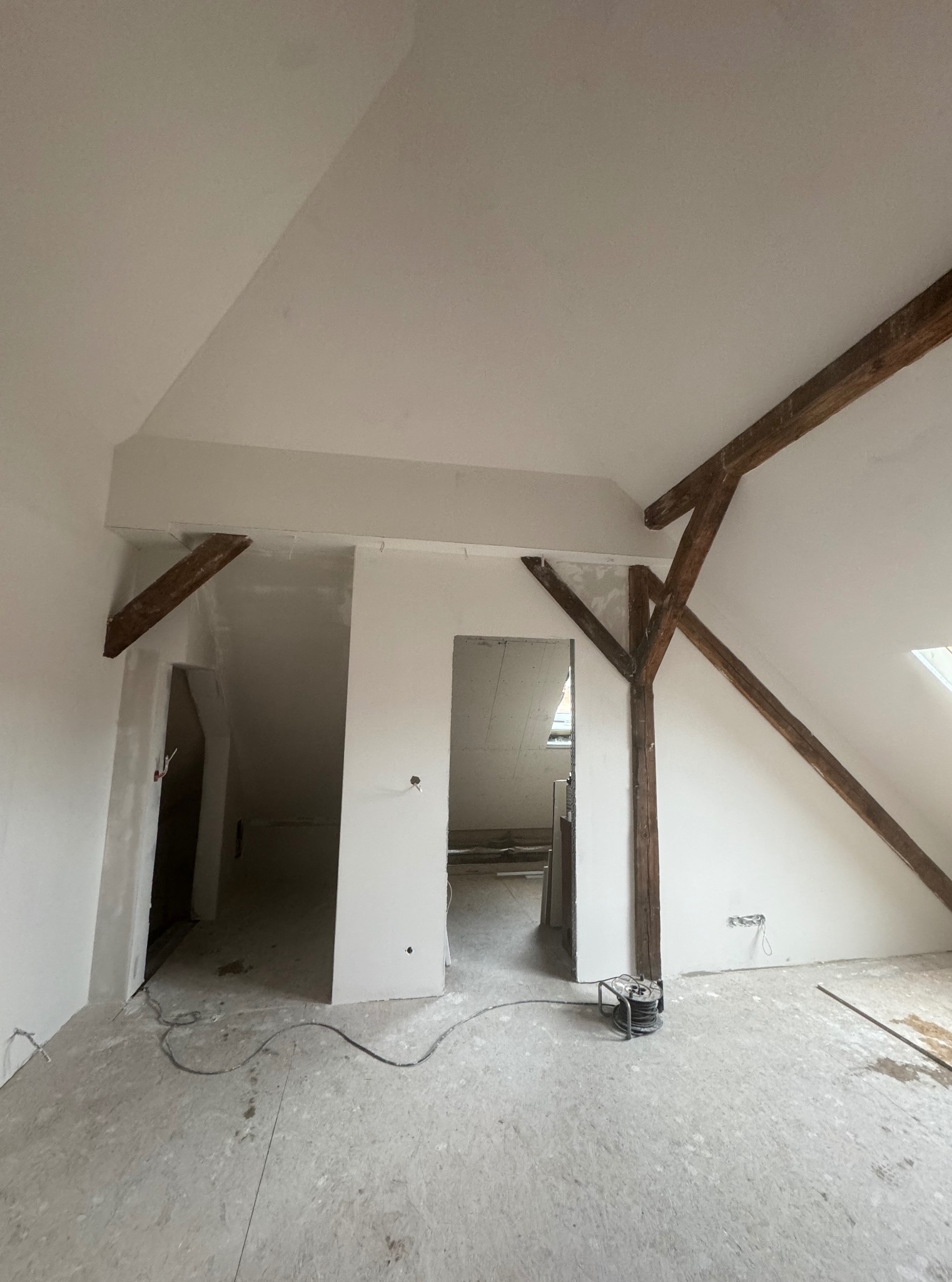 Poddasze po szpachlowaniu, widoczne drewniane elementy konstrukcyjne, ściany przygotowane do malowania, podłoga zabezpieczona folią. Widok z dołu, szeroki kąt.