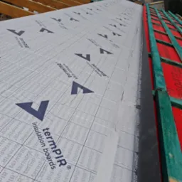 Montaż srebrnej folii termoizolacyjnej z nadrukiem na drewnianej konstrukcji dachu, widoczne fragmenty łat i kontrłat.