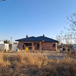 Tab-Dach - Niedokończony dom jednorodzinny z ciemnym pokryciem na dachu, widoczny zza wysokiej trawy, w tle błękitne niebo i słupy energetyczne.