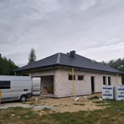 Nowy, grafitowy dach z kominem na parterowym domu w trakcie budowy, widoczne bloczki SOLBET, szara furgonetka i rynny w kolorze żółtym.