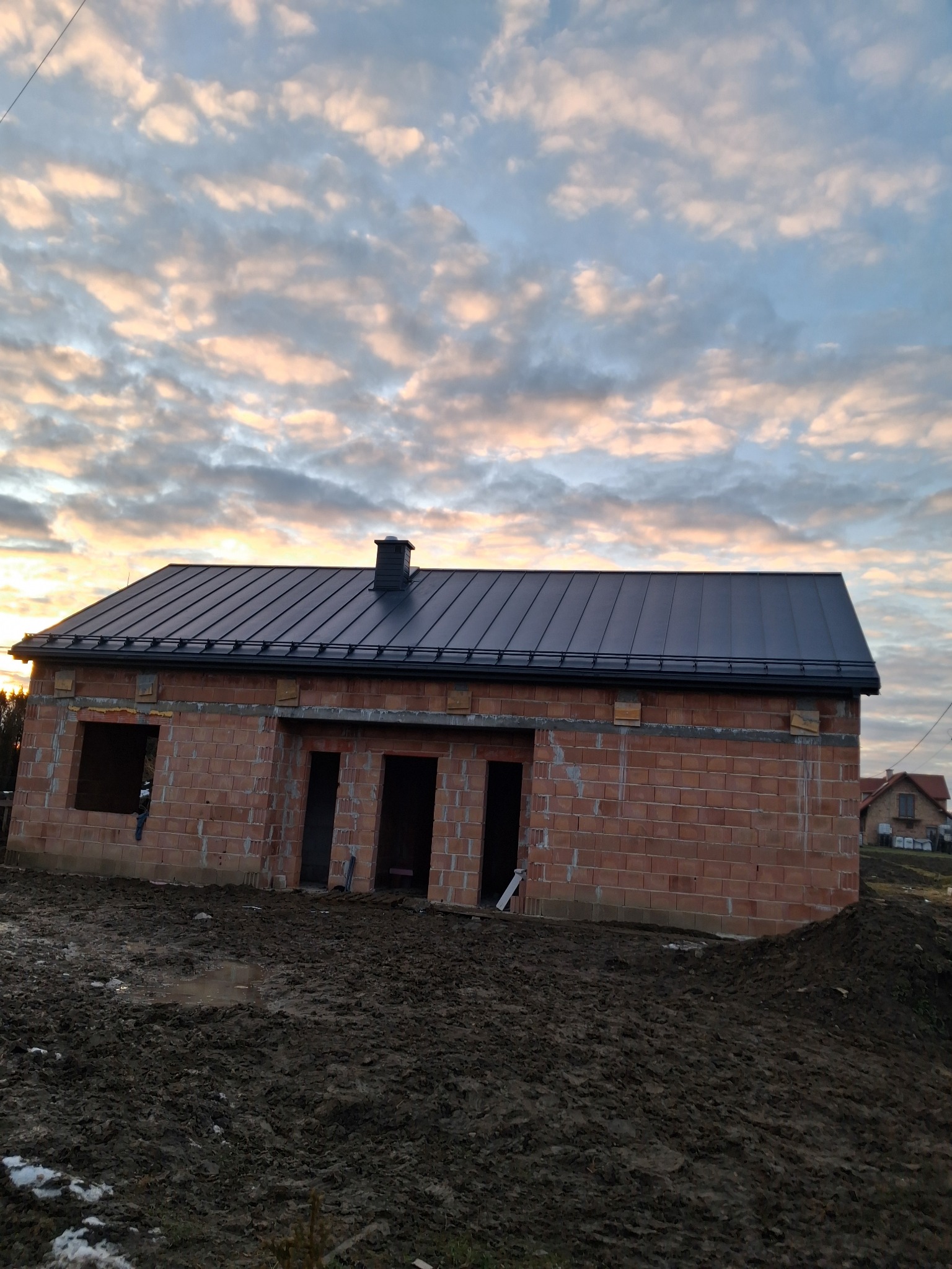 Surowa bryła domu z czerwonej cegły, z zamontowanym, ciemnym dachem blaszanym, na tle zachmurzonego nieba o zachodzie słońca, teren wokół domu rozkopany i błotnisty.