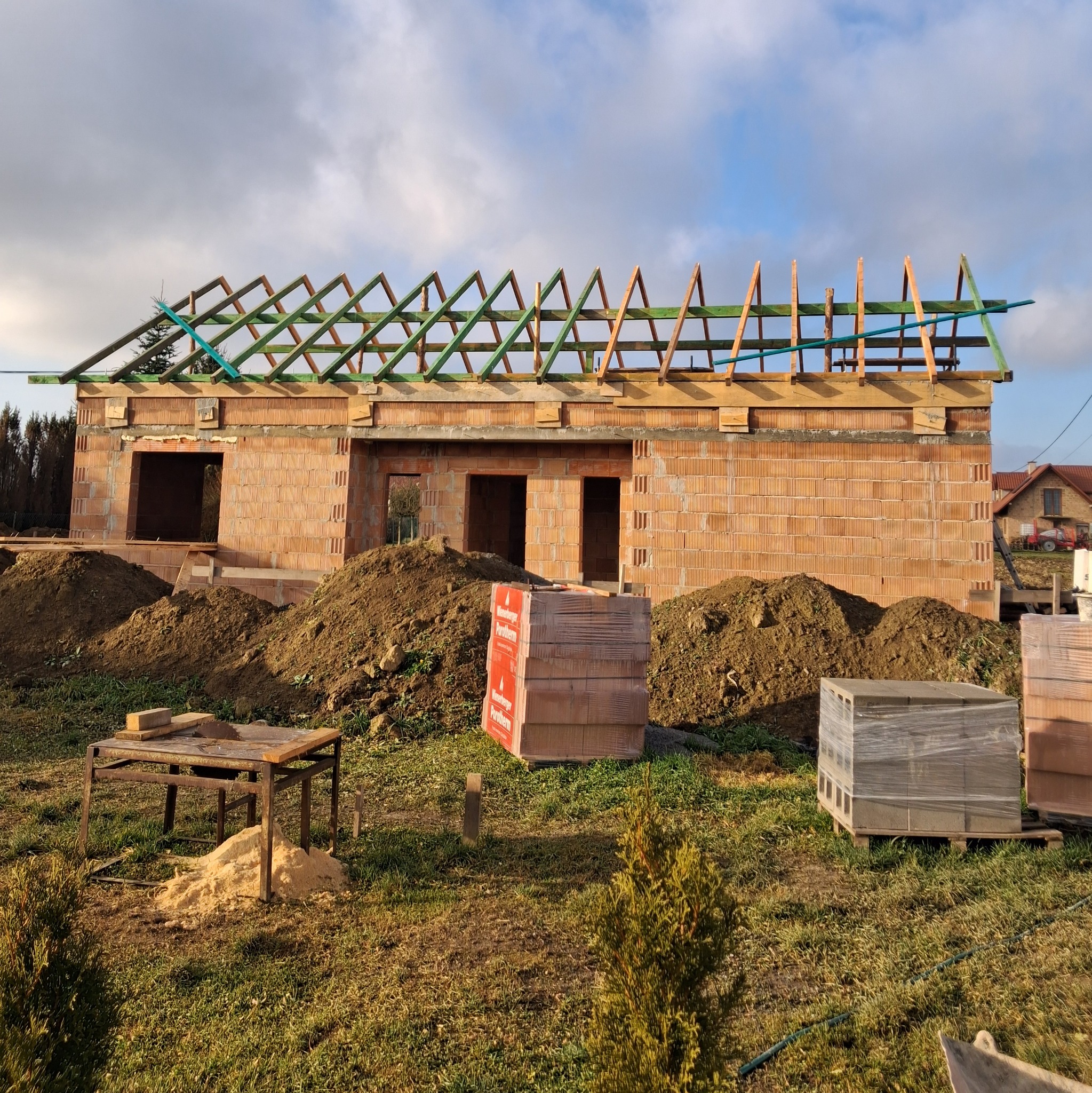 Budowa domu jednorodzinnego w trakcie wznoszenia więźby dachowej, widoczne bloczki ceramiczne Porotherm, sterta ziemi i stół z piłą do drewna.