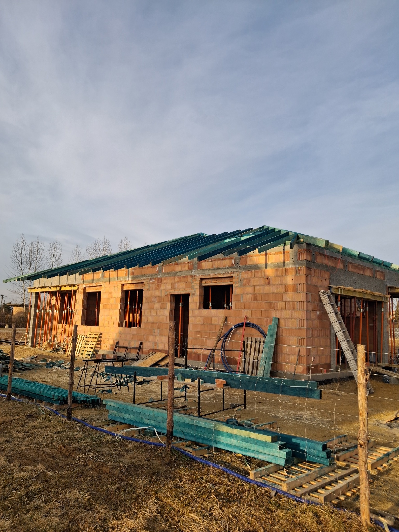 Surowy stan budynku jednorodzinnego z częściowo wykonaną konstrukcją dachu z zieloną impregnacją, widoczne pustaki ceramiczne i przygotowane drewniane elementy więźby dachowej, drabina oparta...