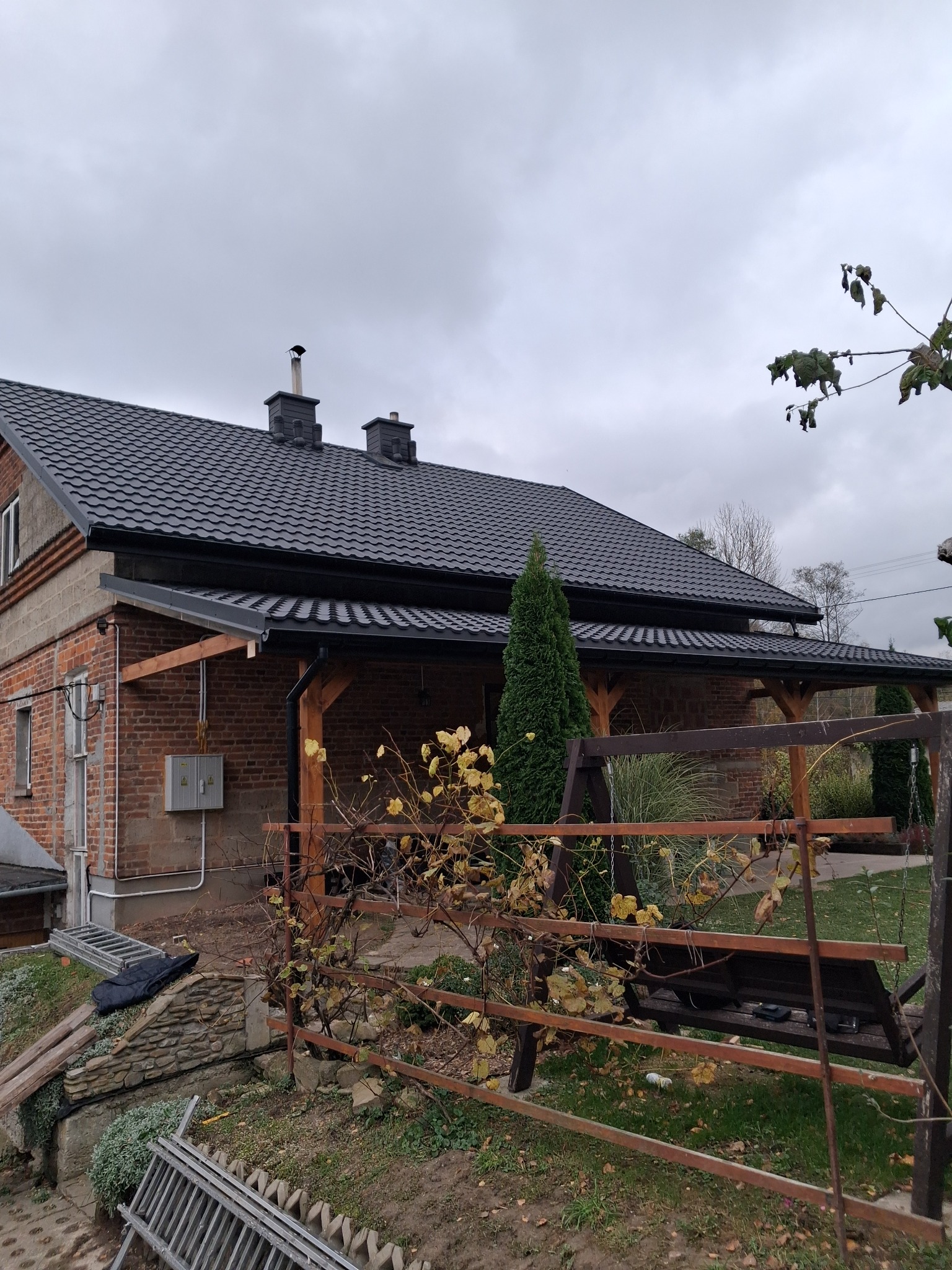 Dom jednorodzinny z ciemnoszarą blachodachówką, widoczne dwa kominy, drewniana konstrukcja zadaszenia tarasu i metalowa drabina oparta o mur.