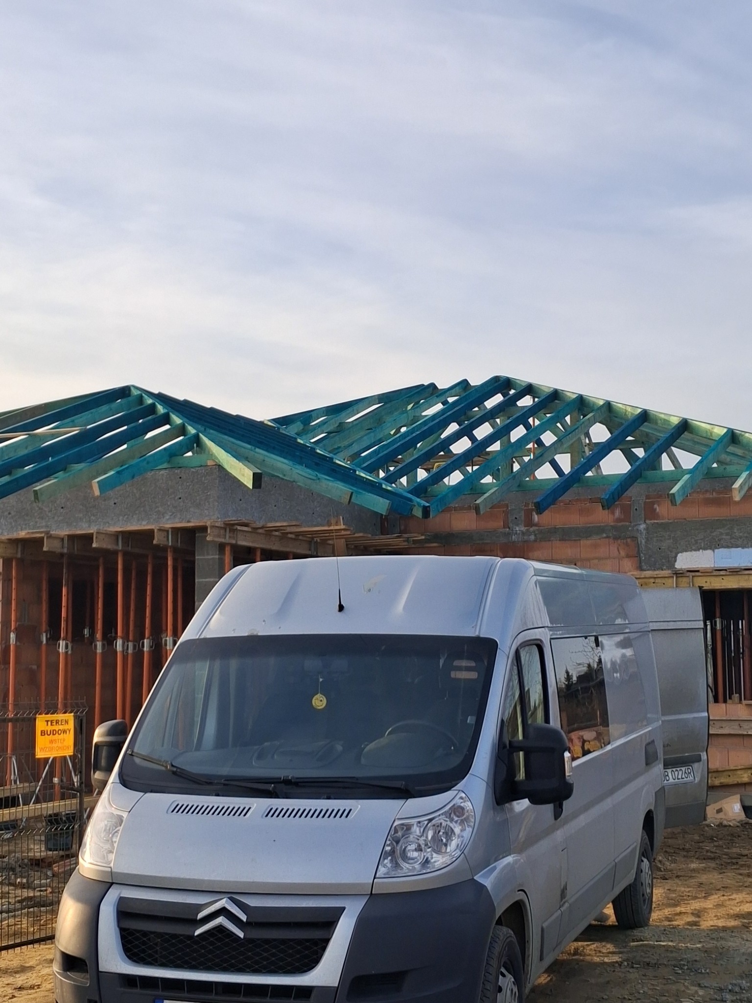 Srebrny furgon Citroen na tle budowy domu z widoczną konstrukcją dachu z impregnowanego na niebiesko drewna i fragmentem murowanych ścian.