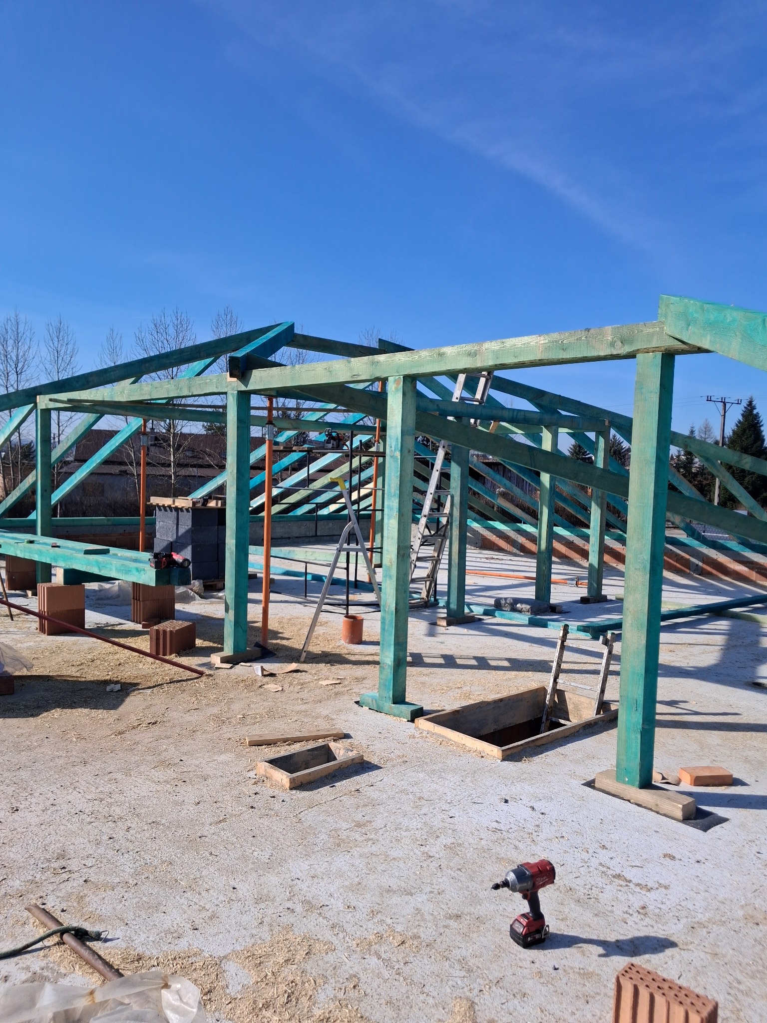 Konstrukcja drewniana dachu w trakcie budowy, pomalowana na zielono-turkusowy kolor, widoczne cegły, drabina i wkrętarka akumulatorowa na tle błękitnego nieba.