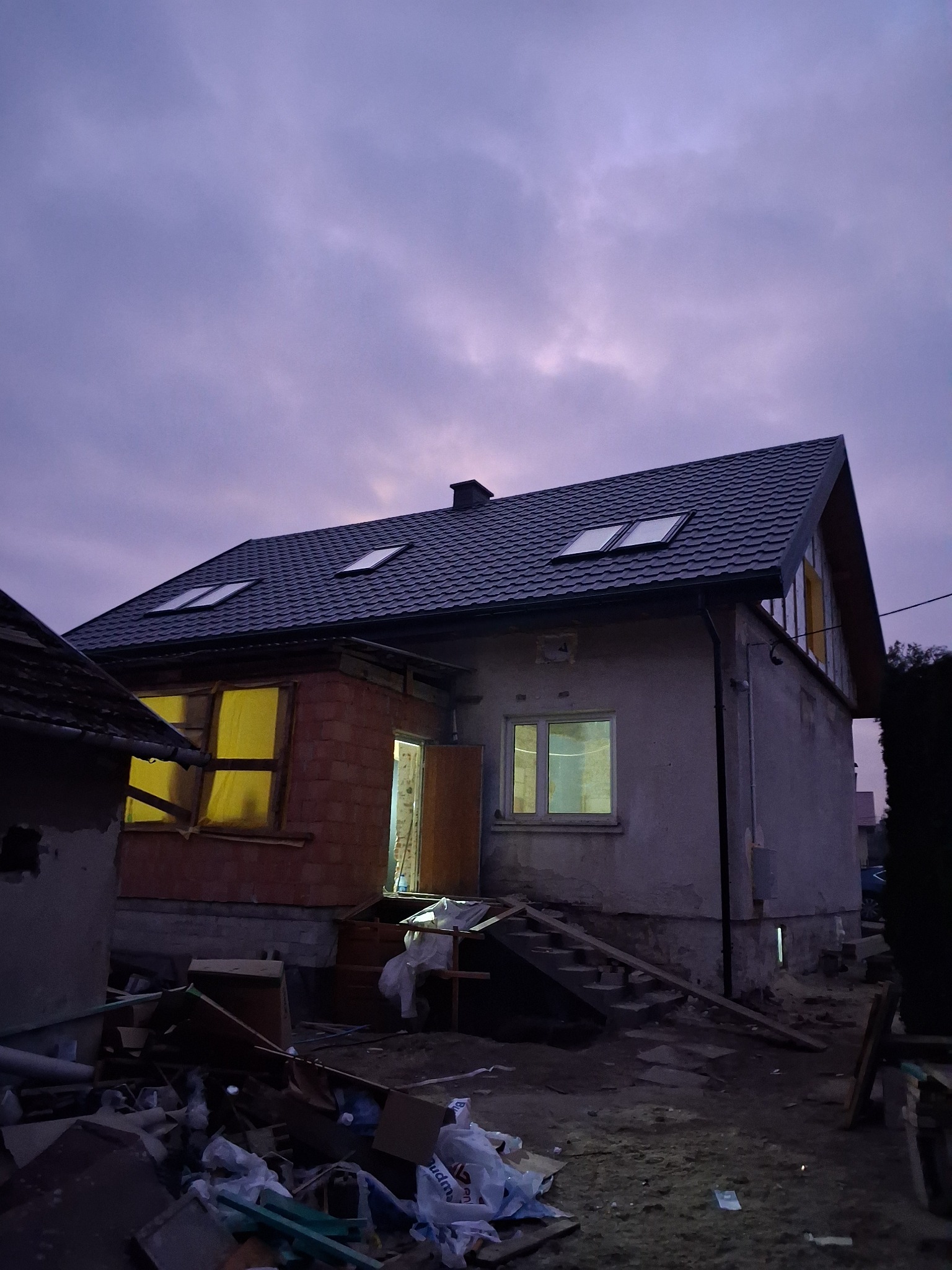 Dom w trakcie budowy z widocznym nowym pokryciem dachowym, oknami dachowymi i dobudowaną częścią z czerwonej cegły. Na podwórku widoczny bałagan budowlany.