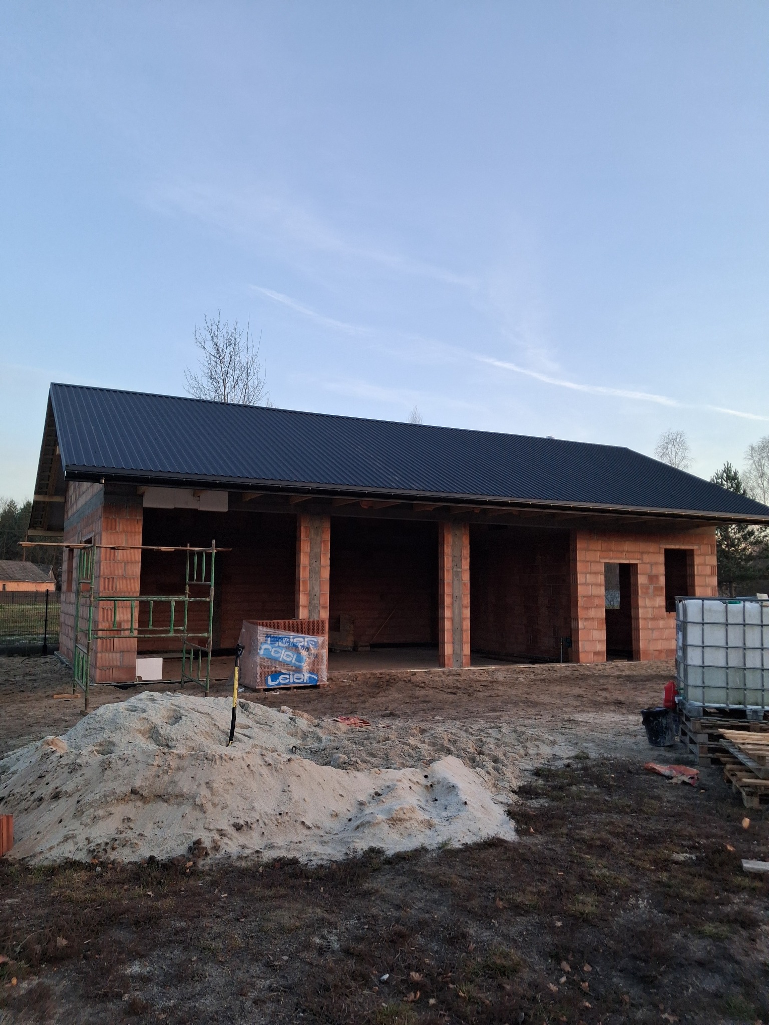 Surowy budynek w trakcie budowy z ciemnym, metalowym pokryciem. Widoczne rusztowania, palety, piasek i zbiornik na wodę. Murowane ściany z otworami okiennymi i drzwiowymi.