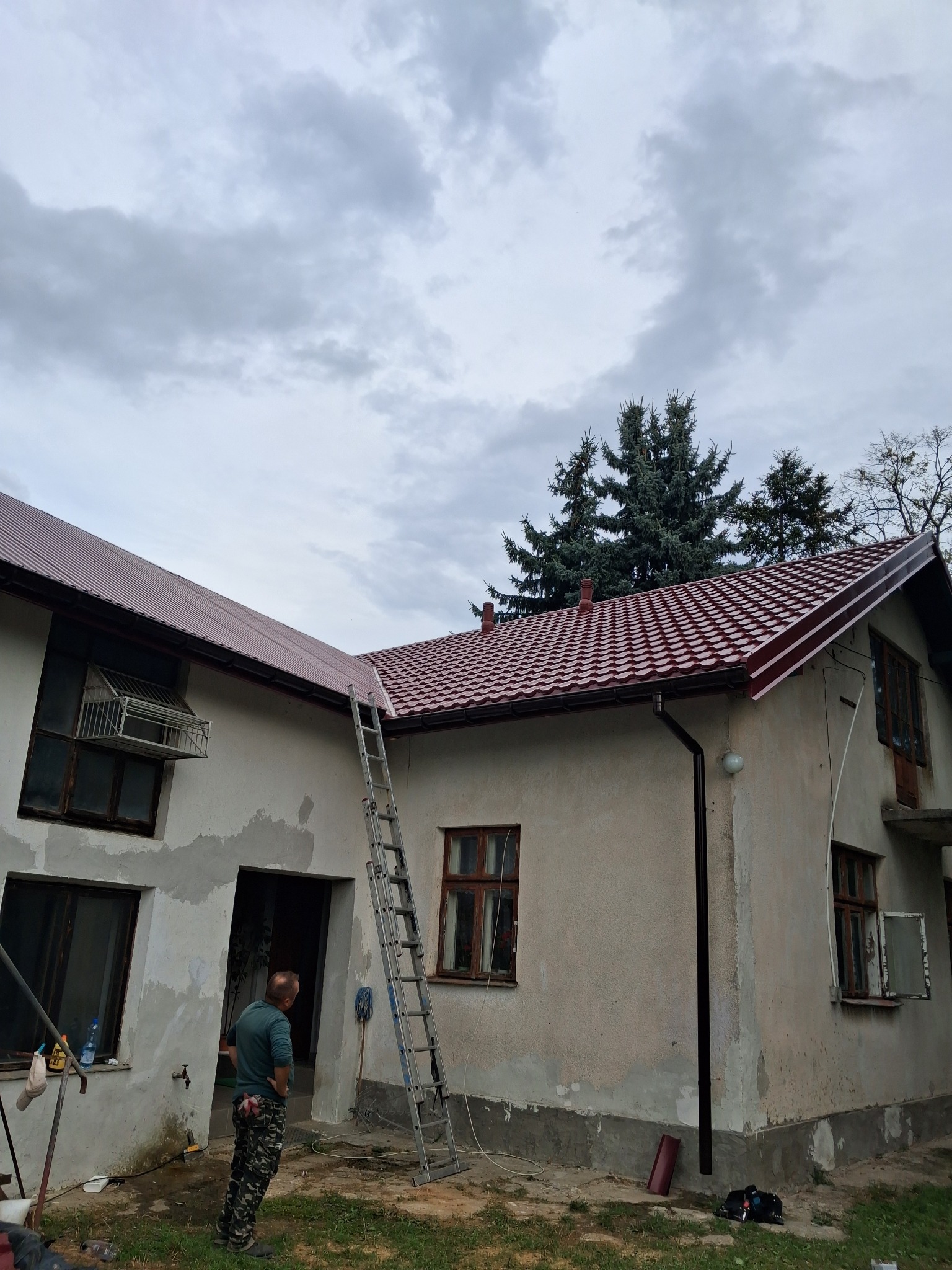 Dom z nowym, czerwonym dachem z blachodachówki, widoczna drabina oparta o ścianę i mężczyzna stojący przed budynkiem.