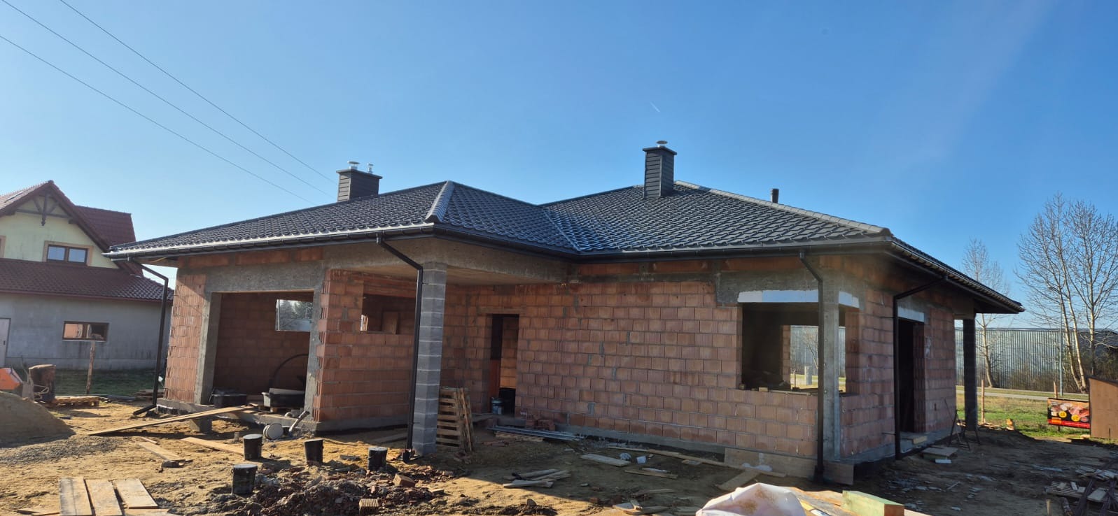 Budynek w trakcie budowy z zamontowanym, ciemnoszarym pokryciem i rynnami, widoczne ceglane ściany i otoczenie placu budowy.