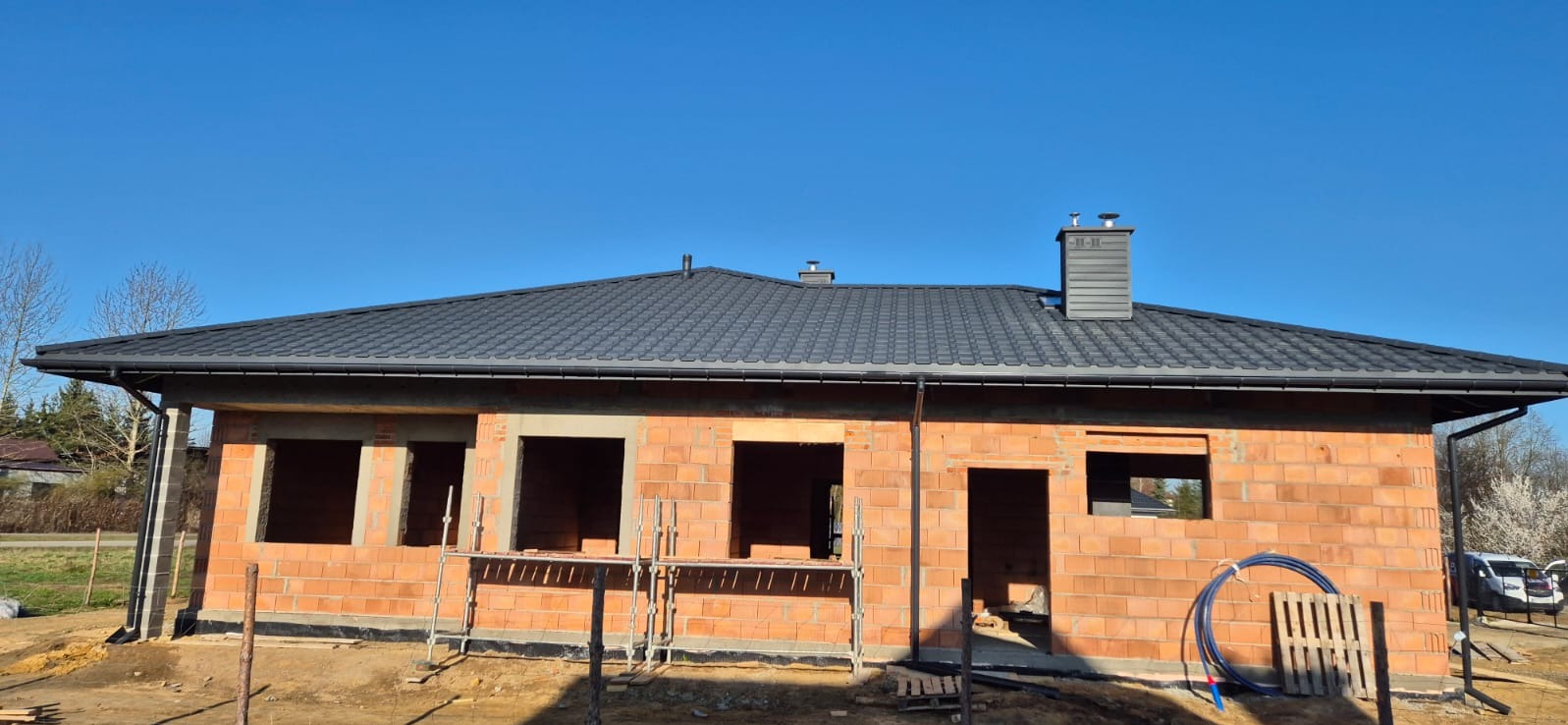 Dom w trakcie budowy z zamontowanym, grafitowym dachem z blachodachówki, widoczne rynny i komin wentylacyjny.