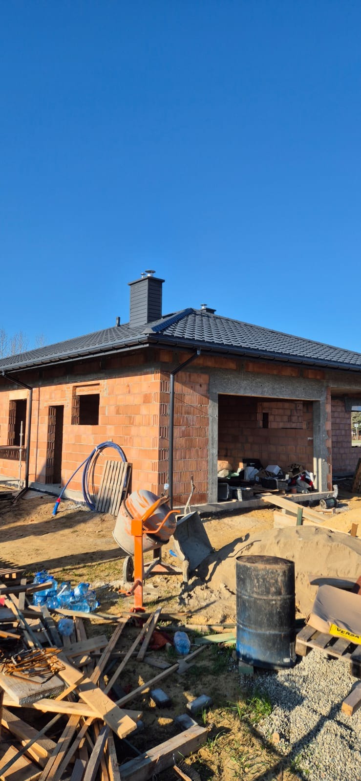 Dom jednorodzinny w trakcie budowy, z widocznym nowym, grafitowym pokryciem, kominem i rynnami. Na pierwszym planie betoniarka, materiały budowlane i śmieci.