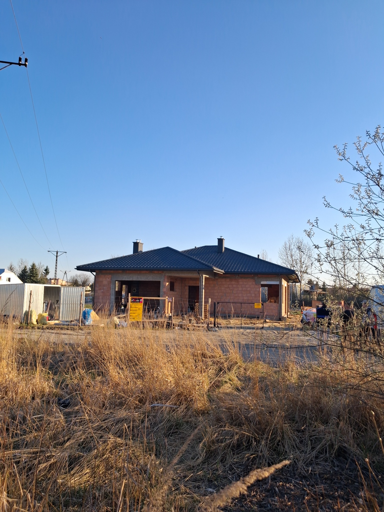 Niedokończony dom jednorodzinny z ciemnym pokryciem na dachu, widoczny zza wysokiej trawy, w tle błękitne niebo i słupy energetyczne.