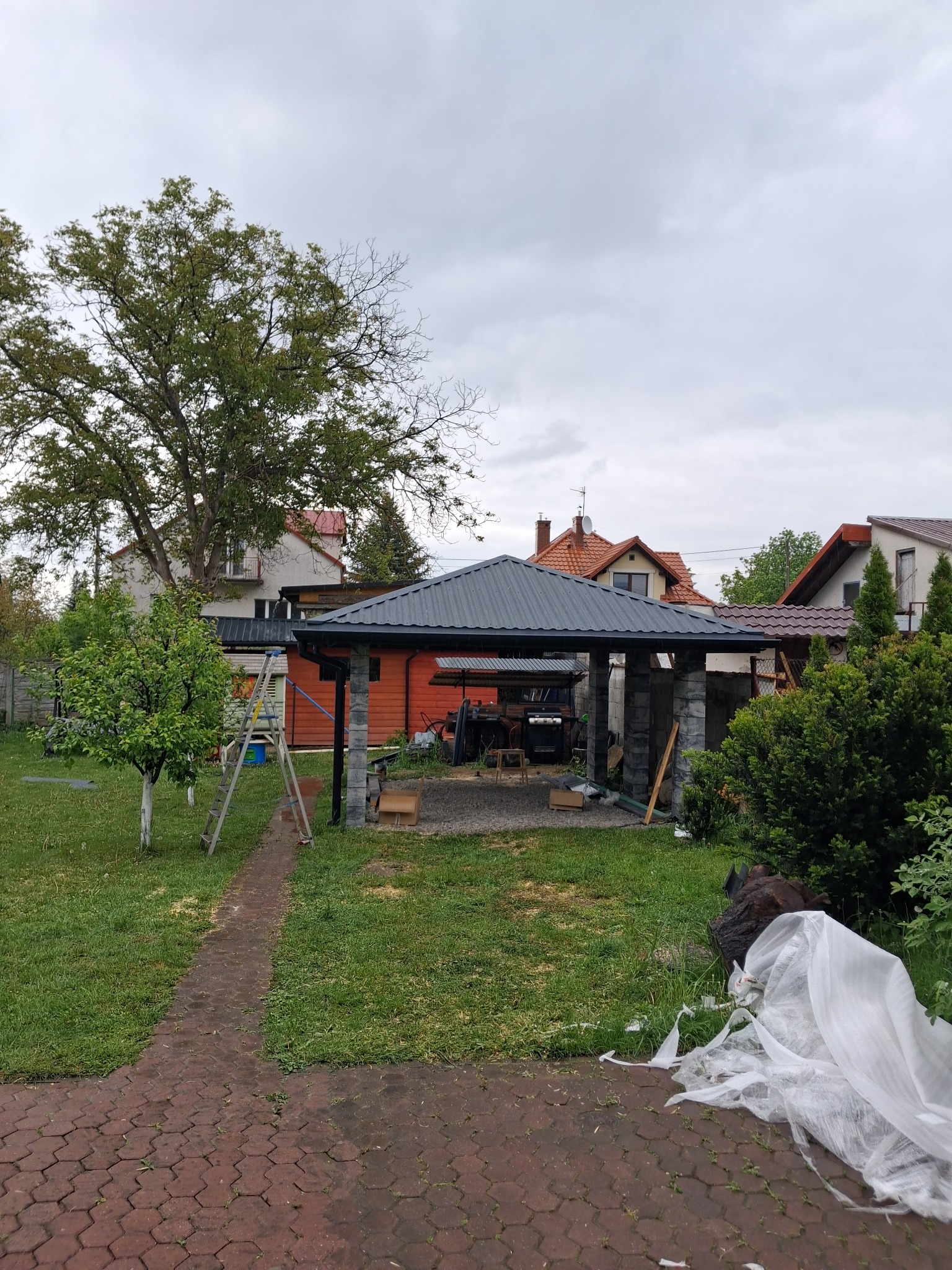 Nowoczesna altana z ciemnym dachem w ogrodzie, w tle domy jednorodzinne i drzewa. Widoczne narzędzia i materiały budowlane, co sugeruje prace wykończeniowe.