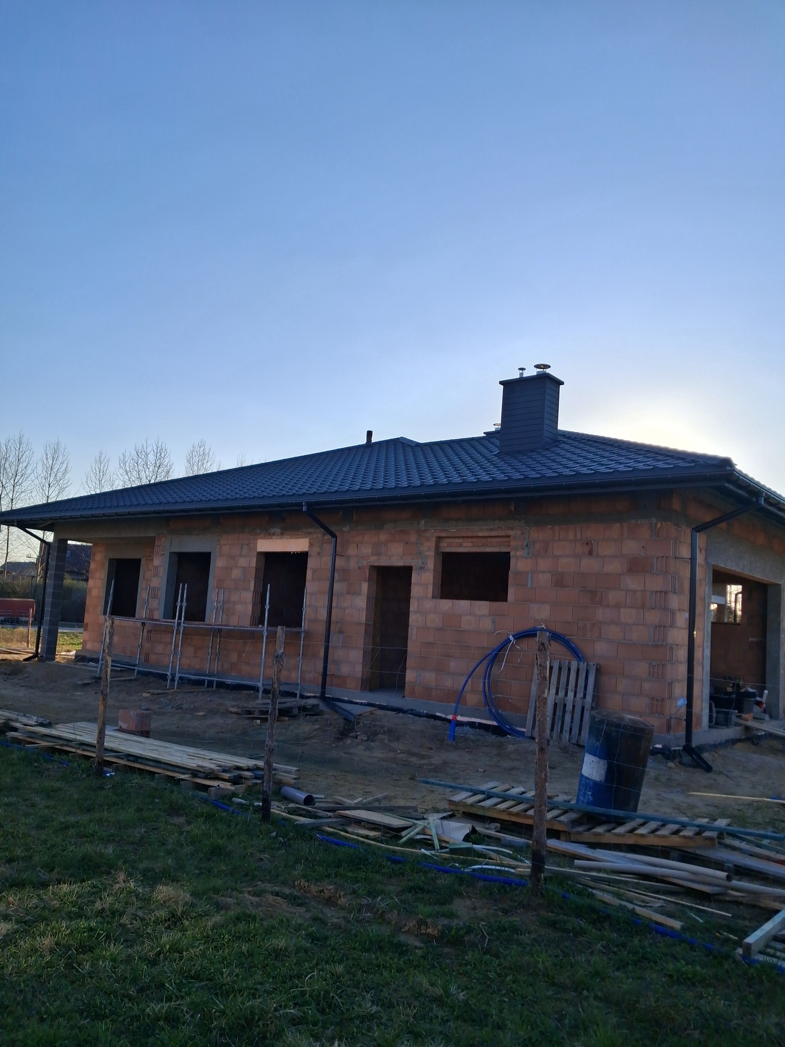 Dom w trakcie budowy z ciemnym dachem i kominem. Widoczne rusztowania, materiały budowlane i trawa. Błękitne niebo w tle.