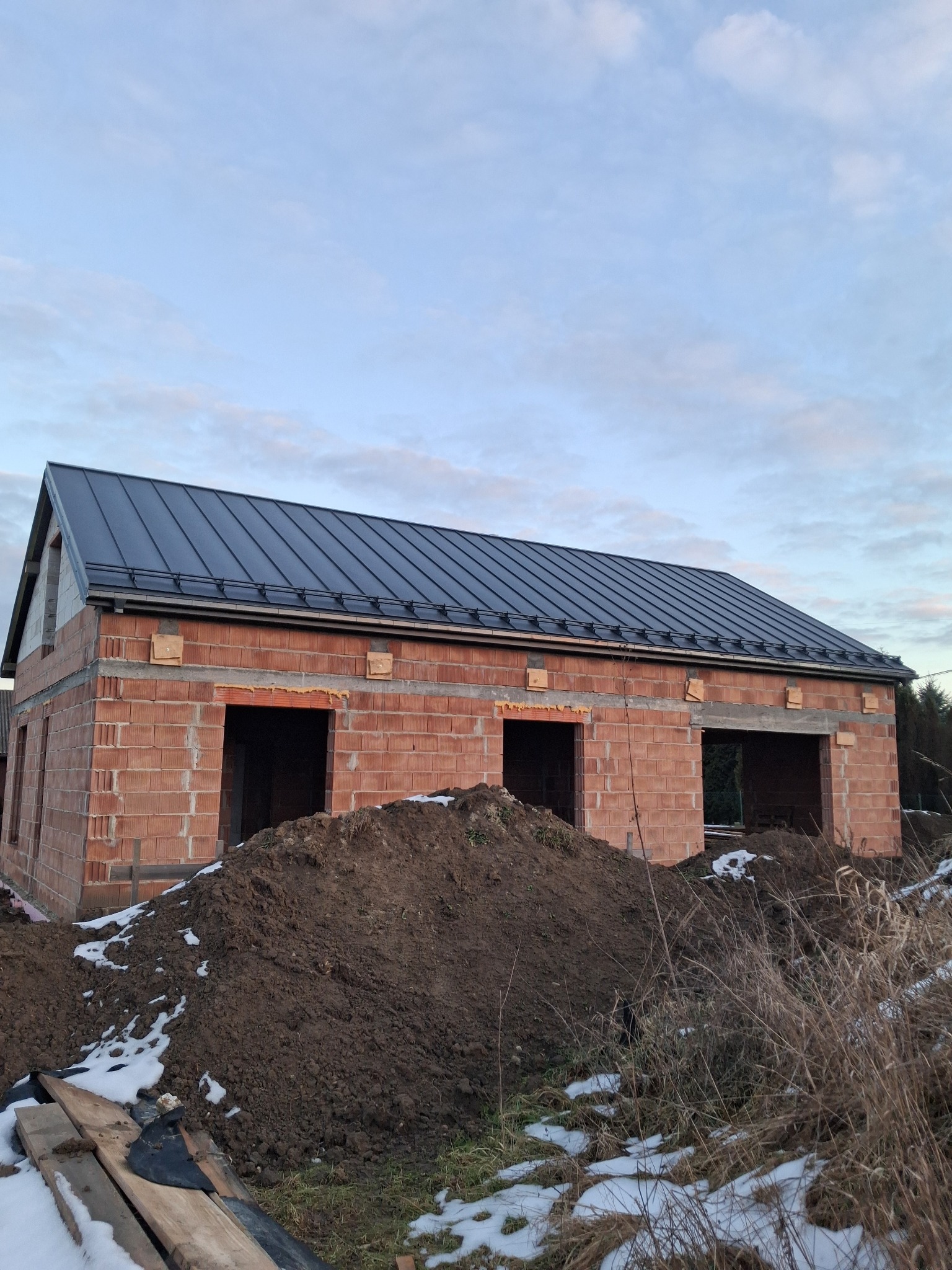 Surowy budynek z czerwonej cegły z ciemnym, nowym dachem blaszanym, widoczne otwory okienne i drzwiowe, sterta ziemi przed budynkiem, zimowa aura.