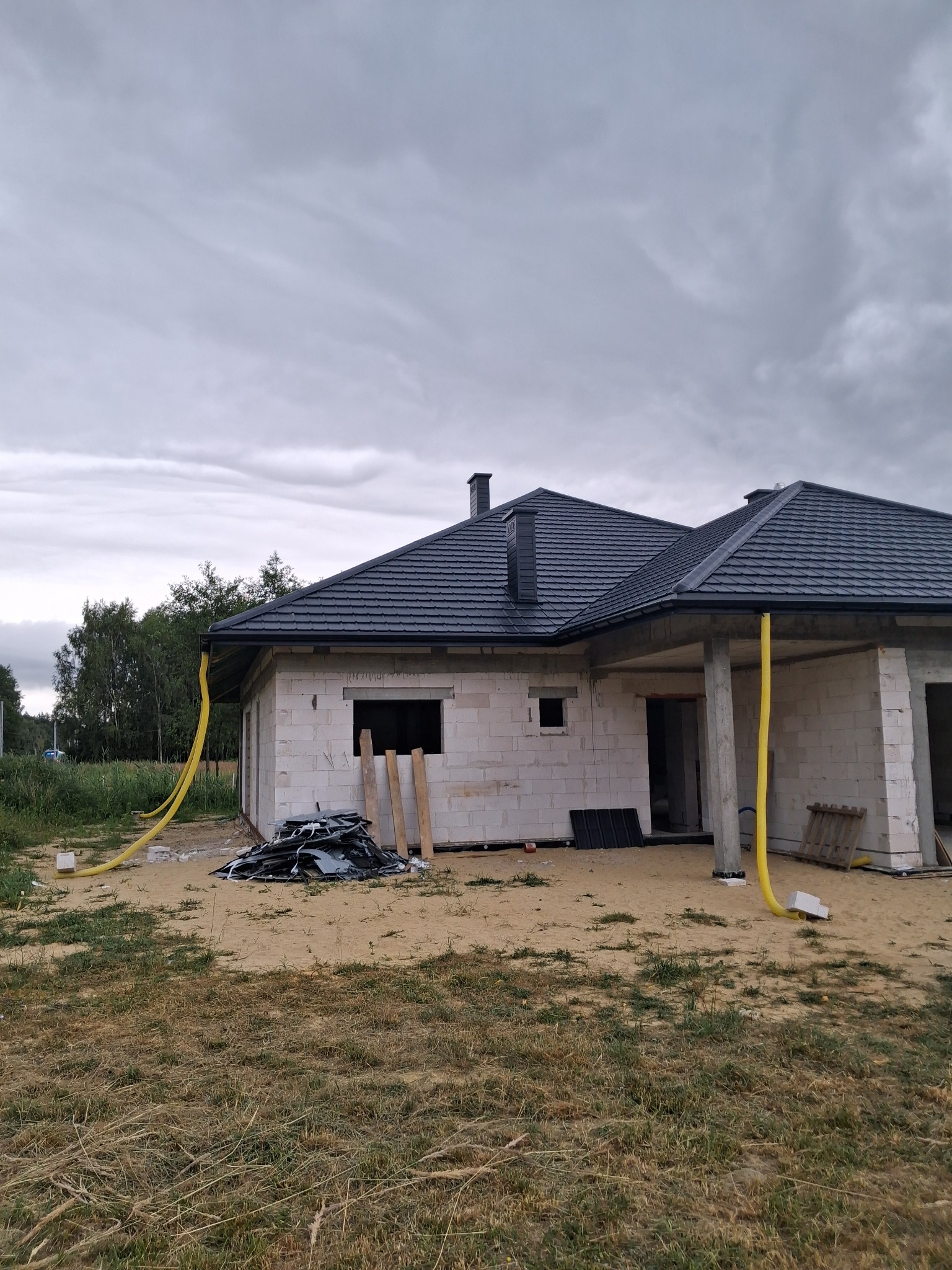 Surowy dom jednorodzinny z ciemnoszarą dachówką karpiówką i żółtymi rurami spustowymi, w trakcie budowy, na tle pochmurnego nieba.