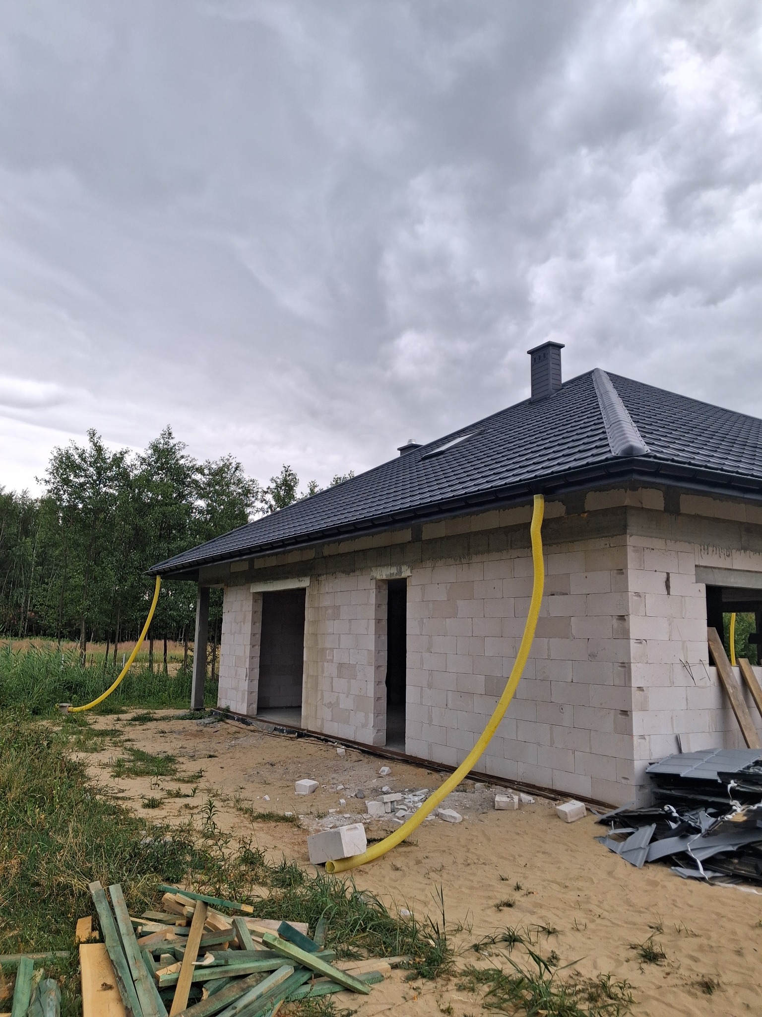 Surowy dom z szarego bloczka, czarny dach z rynnami i żółtymi rurami spustowymi. Na ziemi gruz i drewniane deski. Pochmurne niebo w tle.