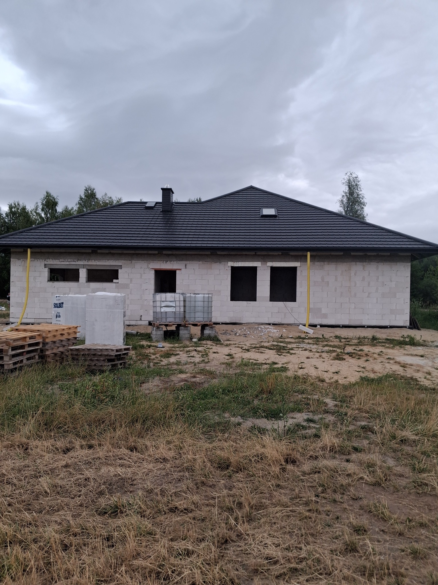 Dom w trakcie budowy z czarnym, skośnym dachem z dachówki ceramicznej. Widoczne palety i materiały budowlane. Pochmurne niebo w tle.
