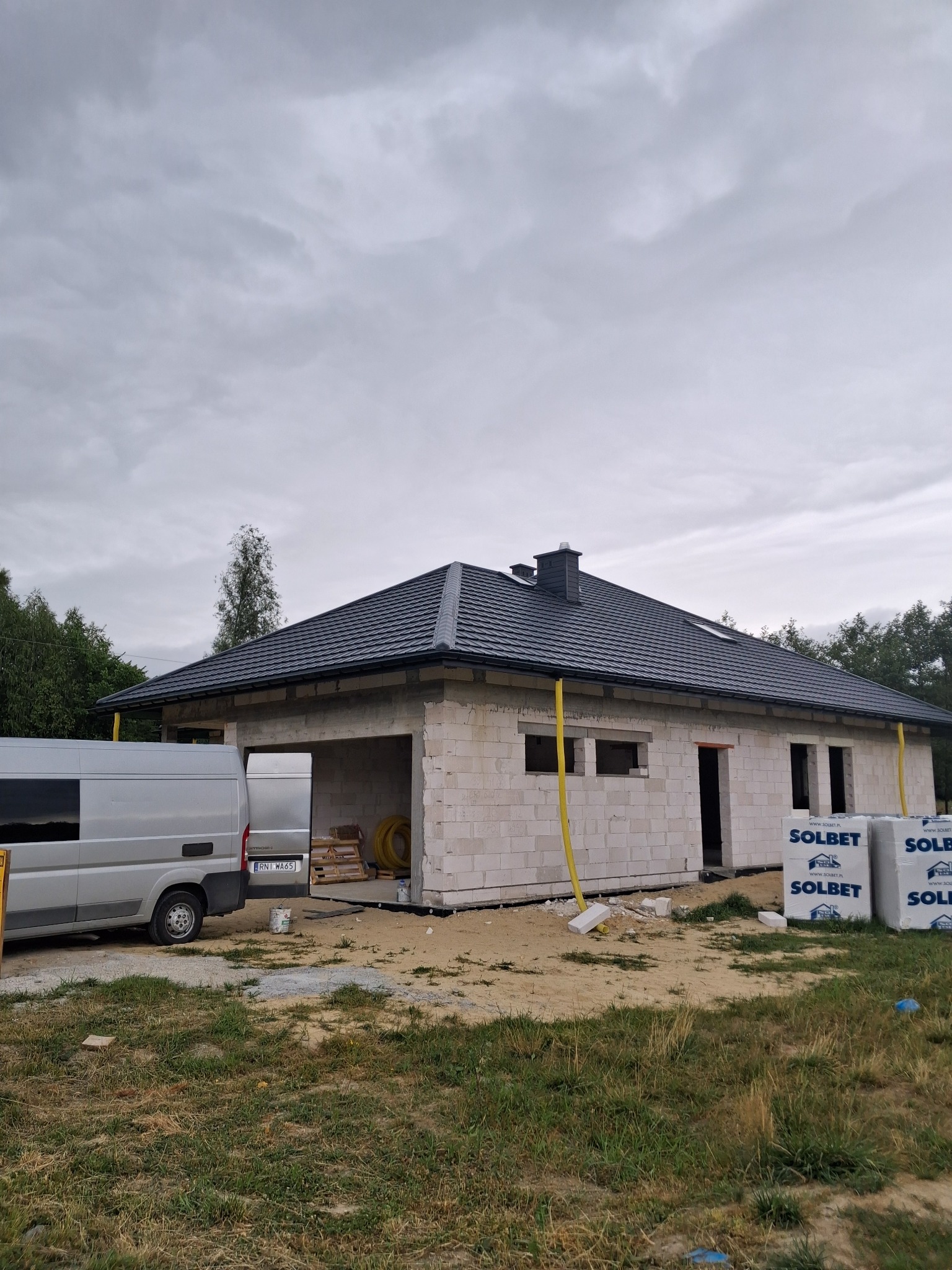 Nowy, grafitowy dach z kominem na parterowym domu w trakcie budowy, widoczne bloczki SOLBET, szara furgonetka i rynny w kolorze żółtym.