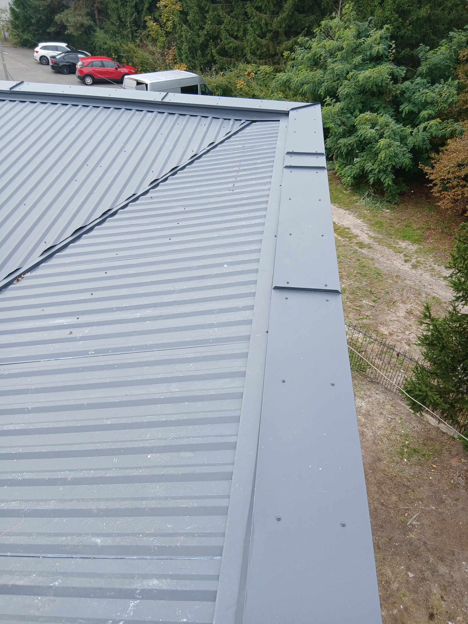 Szary dach z blachy trapezowej, widok z góry, na tle drzew i parkingu. Widoczne obróbki blacharskie na krawędzi dachu, mocowane na śruby.