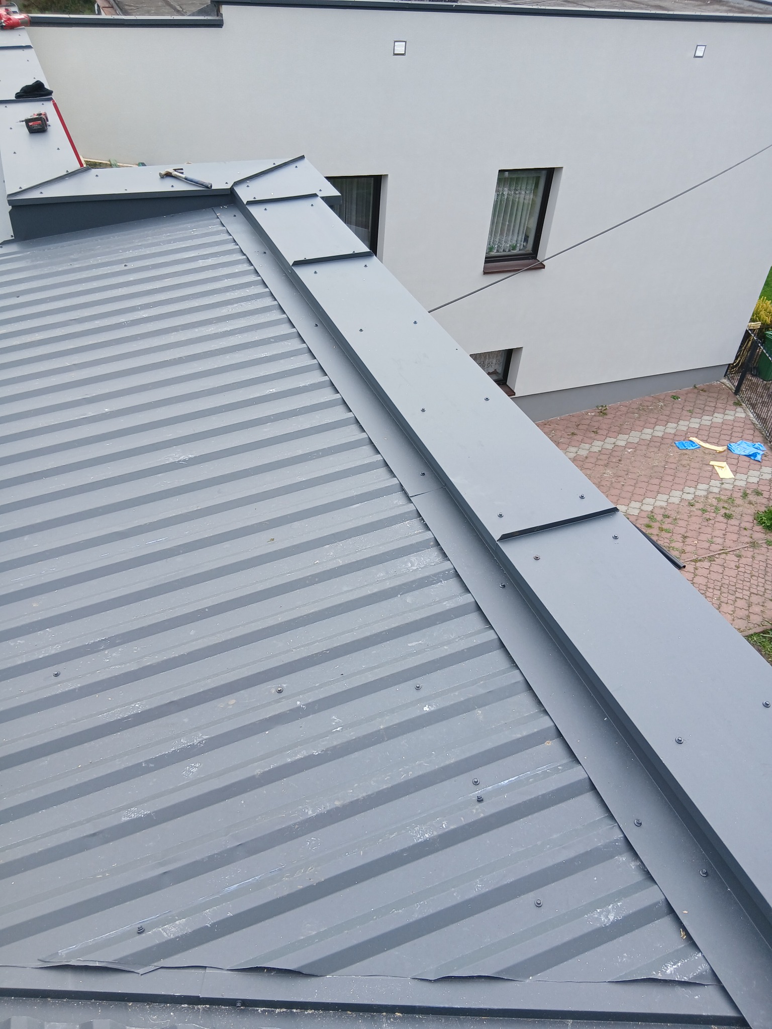 Szary dach z blachy trapezowej z obróbkami blacharskimi na tle białego budynku z oknami i zieleni. Widok z góry, ukośny kadr.