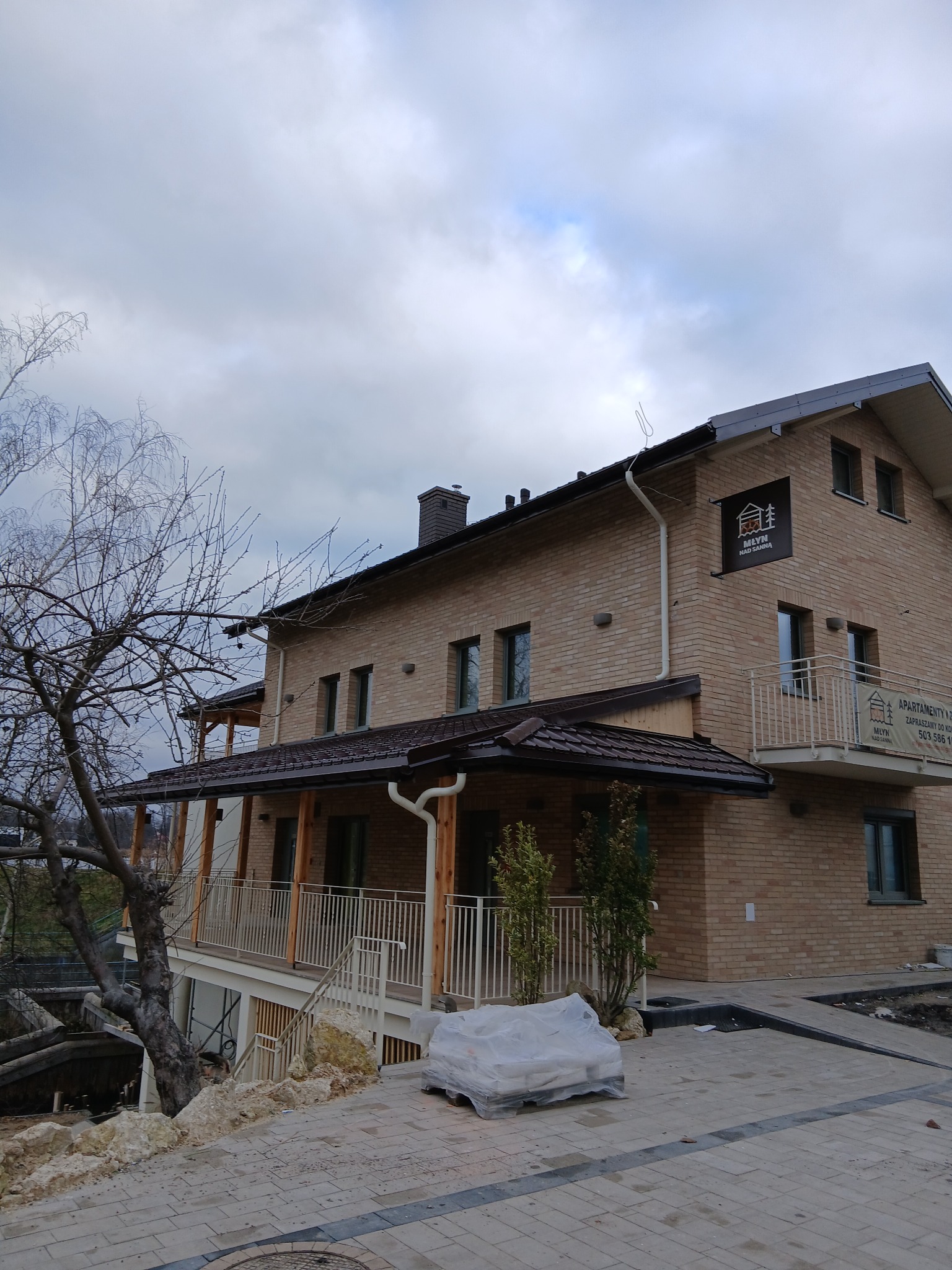 Dwupiętrowy budynek z cegły z brązowym dachem i białymi rynnami, widoczny z dołu pod kątem. Na ścianie logo firmy. Częściowo widoczny balkon z białą balustradą.