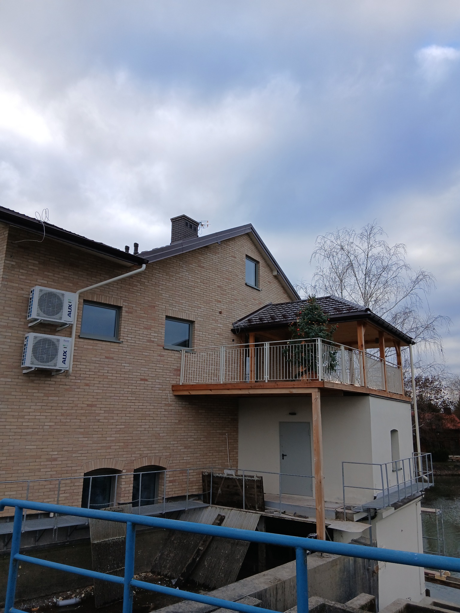 Dwukondygnacyjny budynek z cegły z balkonem i zadaszeniem, widok z dołu, pochmurne niebo w tle. Na ścianie zamontowane klimatyzatory.