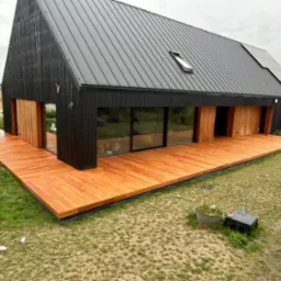 Nowoczesny dom z czarną elewacją i rozległym tarasem z drewna egzotycznego, widok z g&oacute;ry na trawnik z dekoracjami ogrodowymi i bale siana w tle.