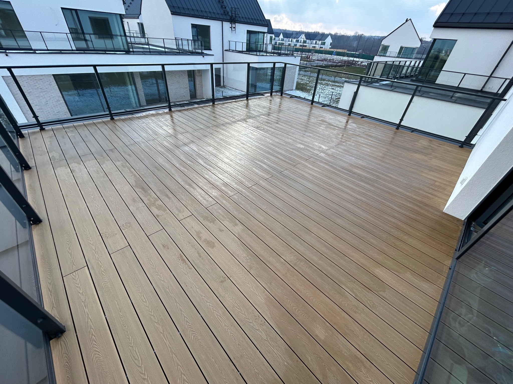 Nowoczesny taras przy apartamentowcu z desek w kolorze jasnego drewna, zabezpieczony szklaną balustradą z czarnymi elementami, widok na inne budynki i fragmenty zieleni w tle.