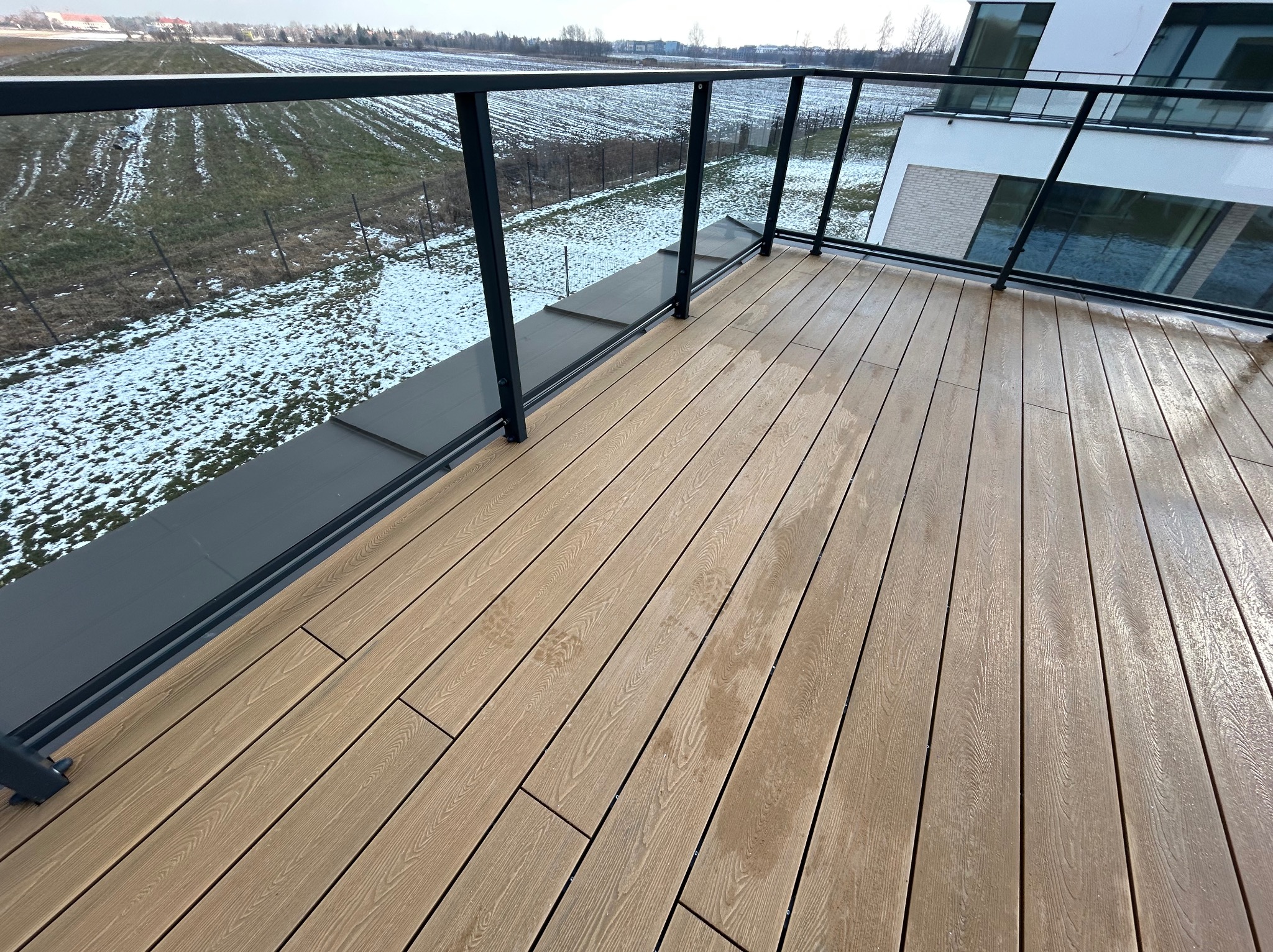 Jasny taras z desek kompozytowych z czarną balustradą szklaną, widok na pole pokryte resztkami śniegu w tle, nowoczesna architektura budynku.