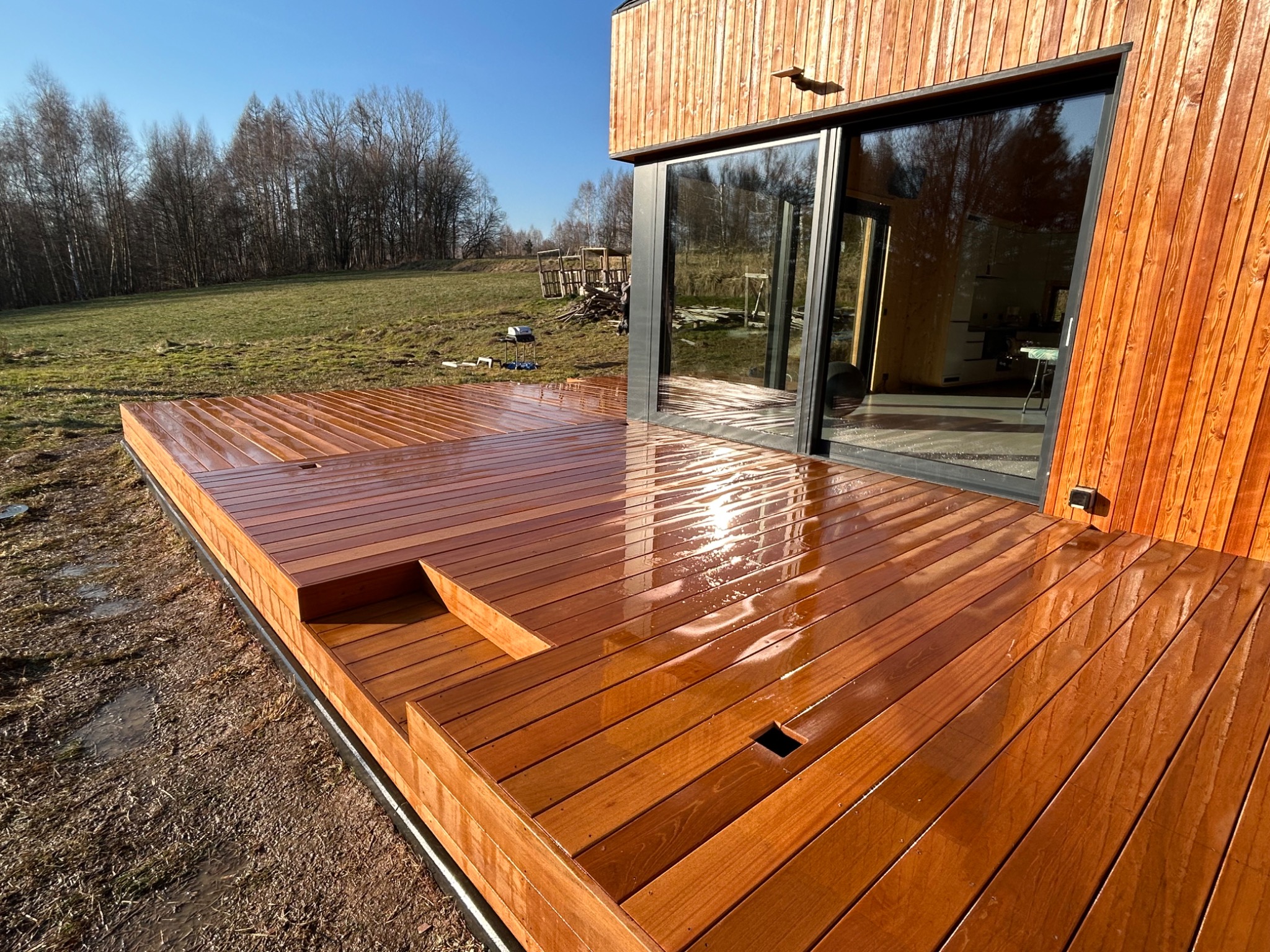 Nowoczesny taras z drewna egzotycznego, po deszczu, z widocznymi śladami wody, przylegający do budynku z drewnianą elewacją i dużymi przesuwnymi drzwiami szklanymi.