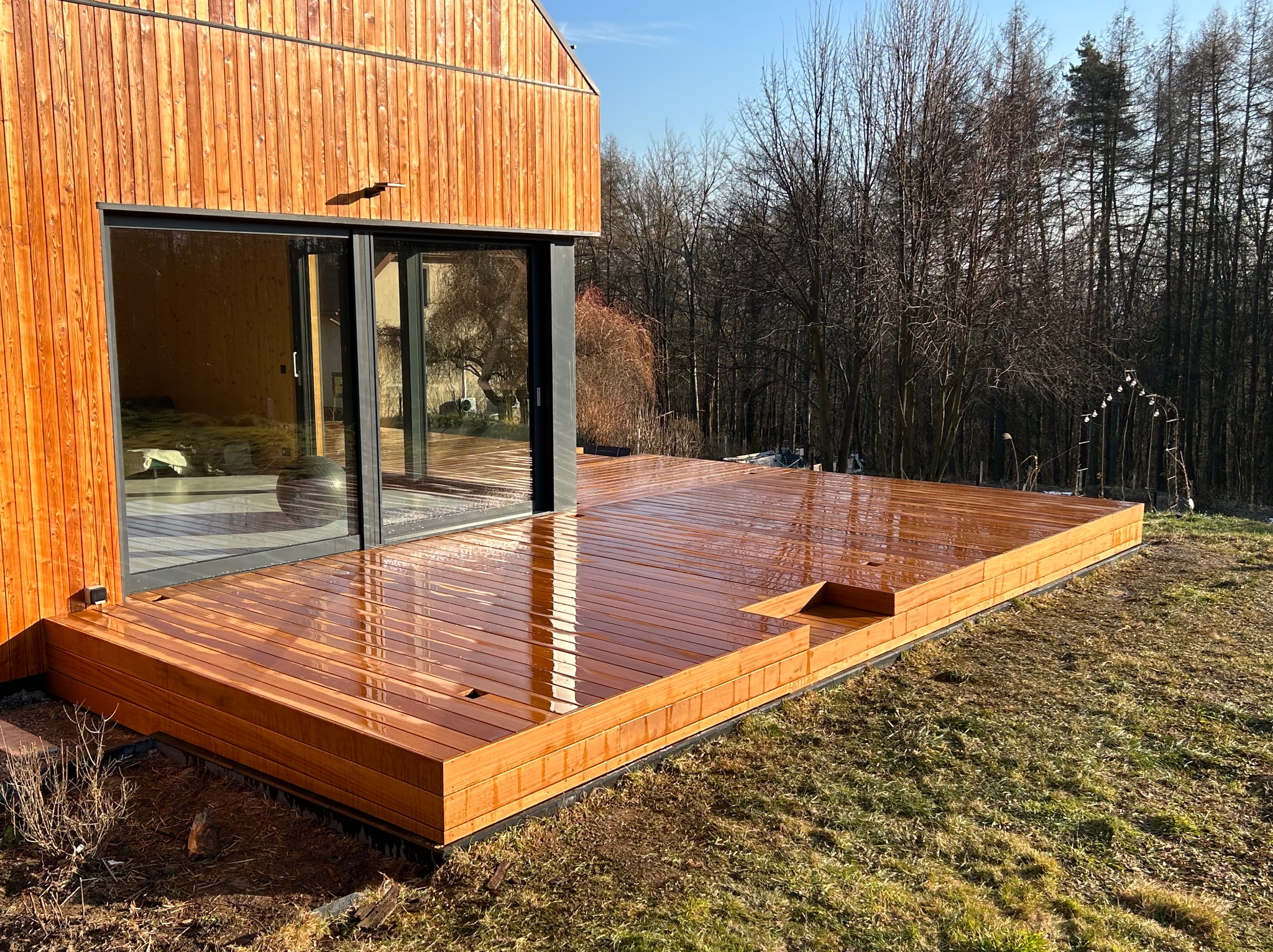 Nowoczesny, drewniany taras przylegający do domu z dużymi, przesuwnymi drzwiami tarasowymi. Deski tarasowe o ciepłym, brązowym odcieniu, ułożone poziomo. W tle widoczny las bezlistny i błękitne niebo.