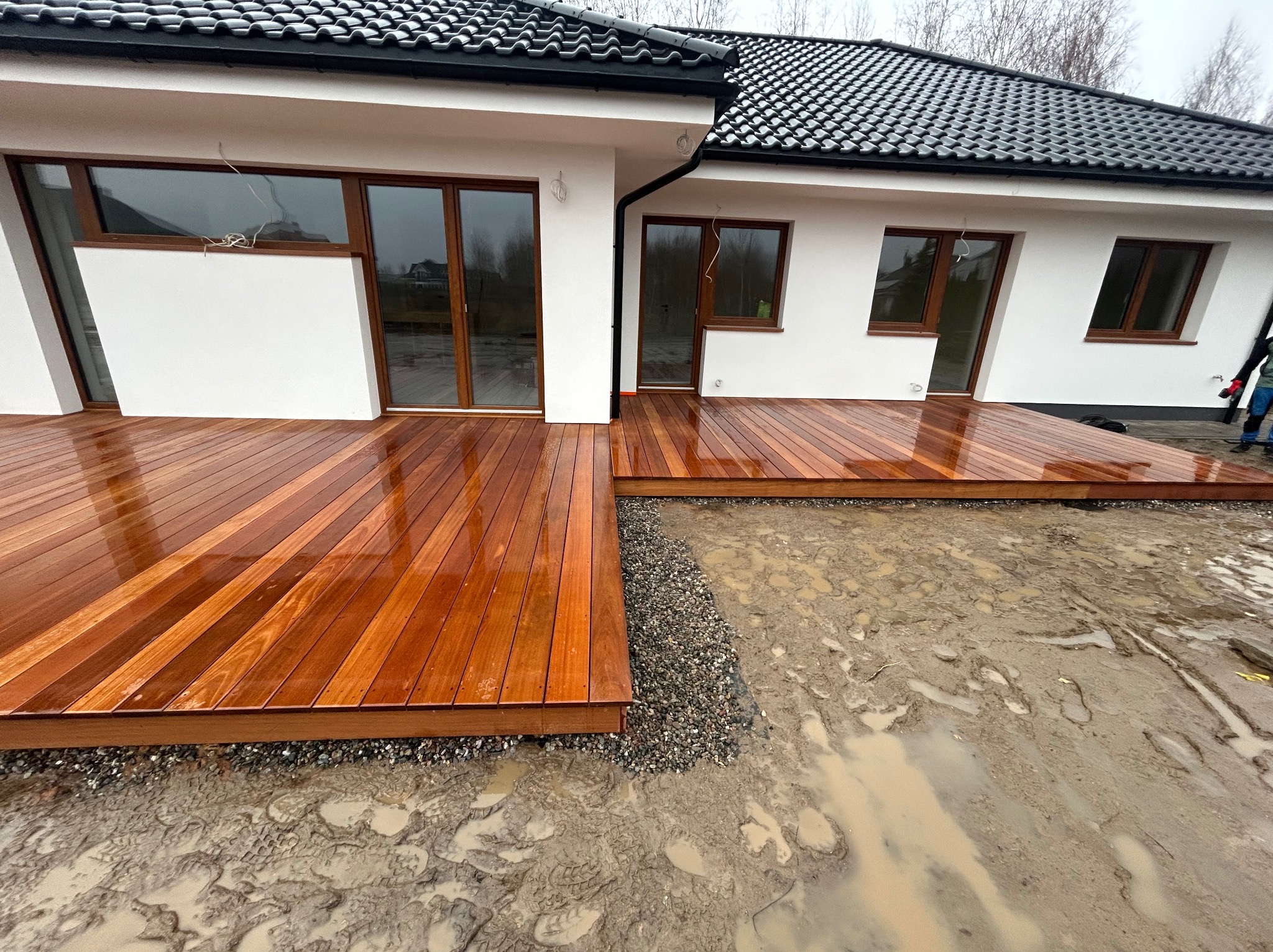Świeżo wybudowany, mokry od deszczu taras z drewna egzotycznego przylegający do białego domu z ciemną dachówką. W tle widoczne błoto i ślady opon.