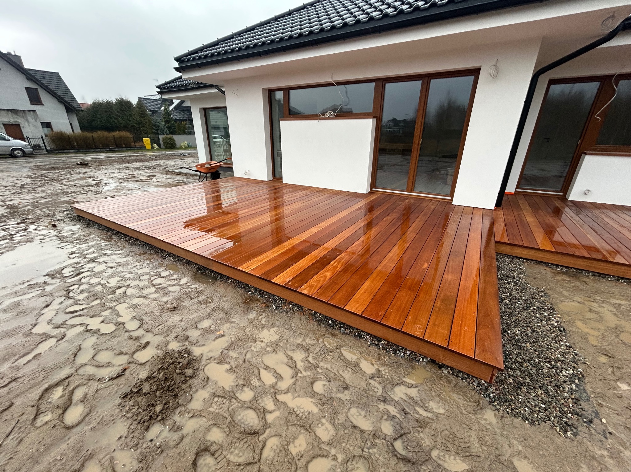 Nowoczesny taras z drewna egzotycznego w Poznaniu, świeżo po deszczu, z widocznymi śladami błota wokół, kontrastujący z minimalistyczną architekturą domu.